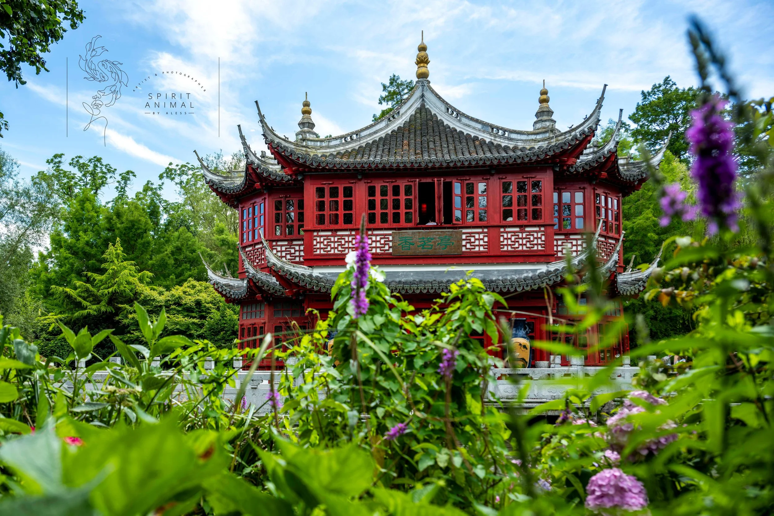 Traditionelles chinesisches Gebäude mit roten Holzstrukturen und doppelstöckigem, geschwungenem Dach, umgeben von grünen Bäumen und bunten Blumen im Vordergrund, unter blauem Himmel, Fotografie von SPIRIT ANIMAL