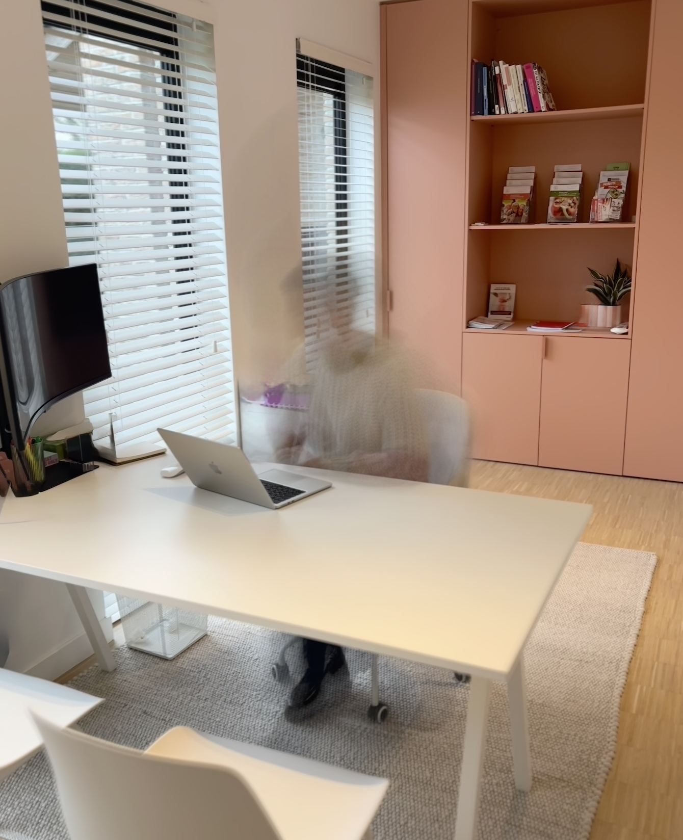 Een kantoorruimte met een witte tafel, een open MacBook, een zwart monitor en diverse kantoorbenodigdheden. Een persoon zit achter de tafel met een lichte trui, beweging is zichtbaar door de beweging op de foto. In de achtergrond bevinden zich grote ramen met witte jaloezieën, een kast met boeken en een plant.