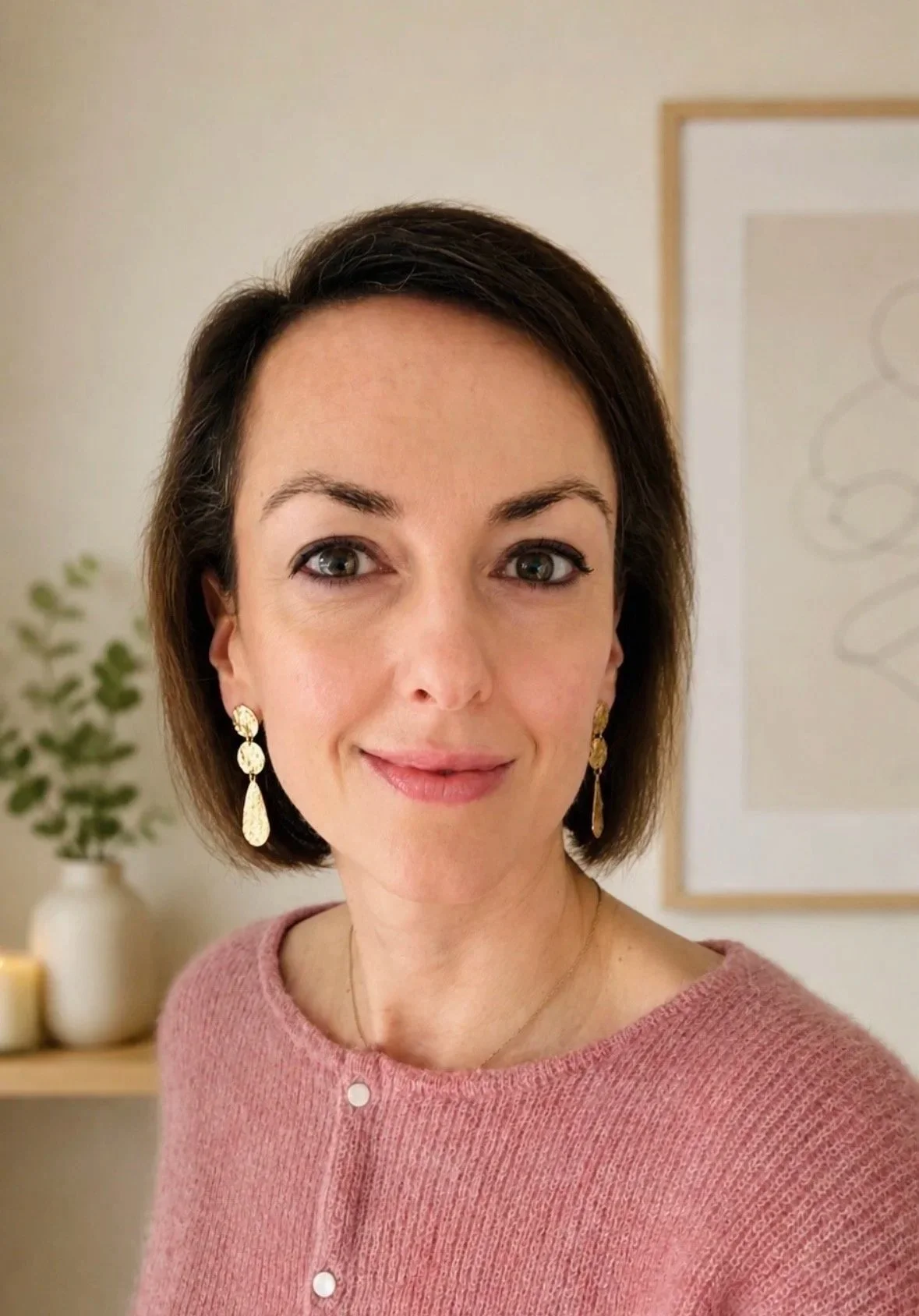 Close-up van een vrouw met korte, donkerbruin haar, die een roze wollen trui en goudkleurige oorbellen draagt, binnen in een kamer met een plant en een kunstwerk op de achtergrond.