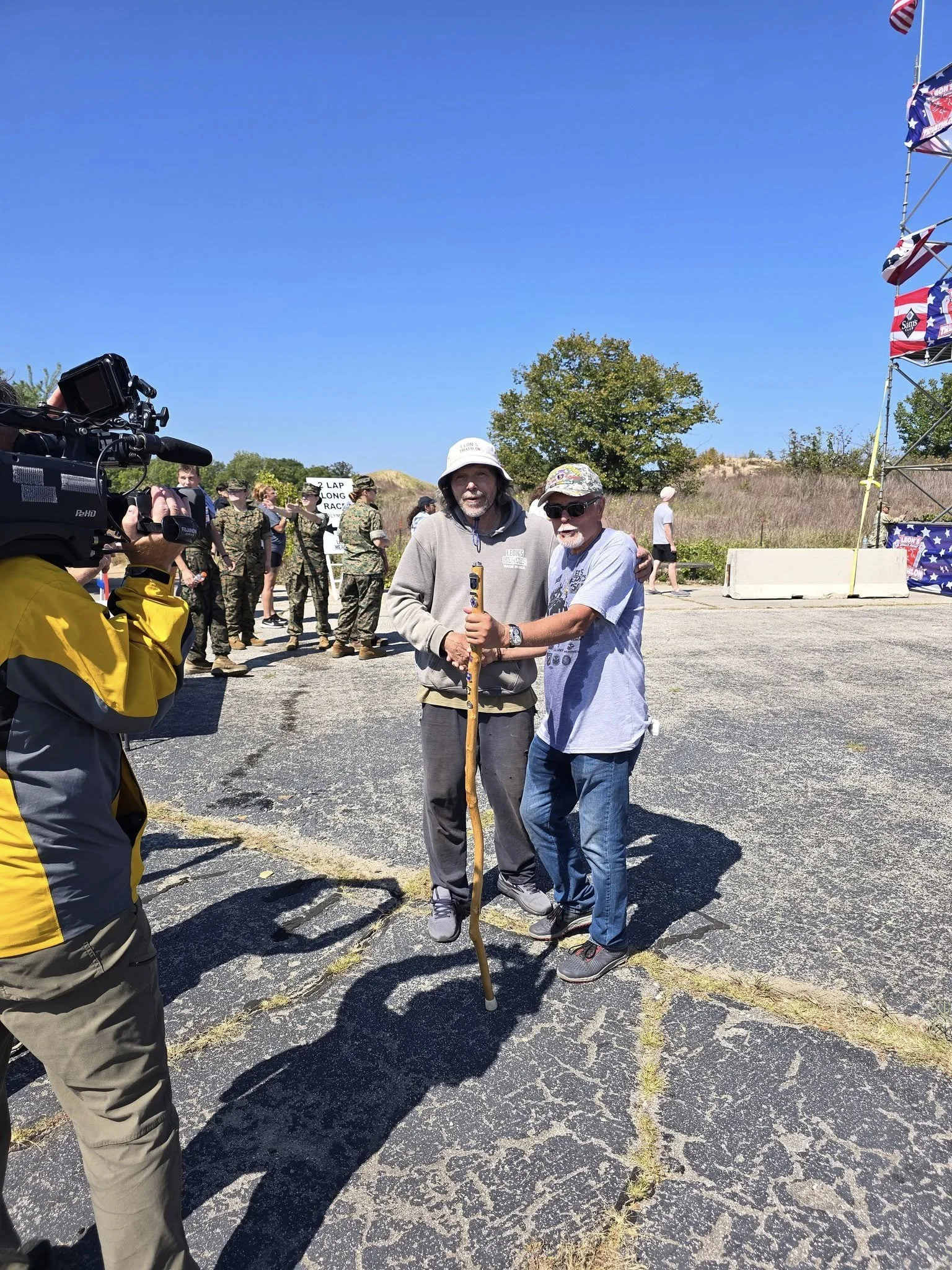 Two men smiling and holding a walking stick together in an outdoor setting, with a camera operator filming them. In the background, there are people in military uniforms, a sign, and a large American flag-themed structure under a clear blue sky.