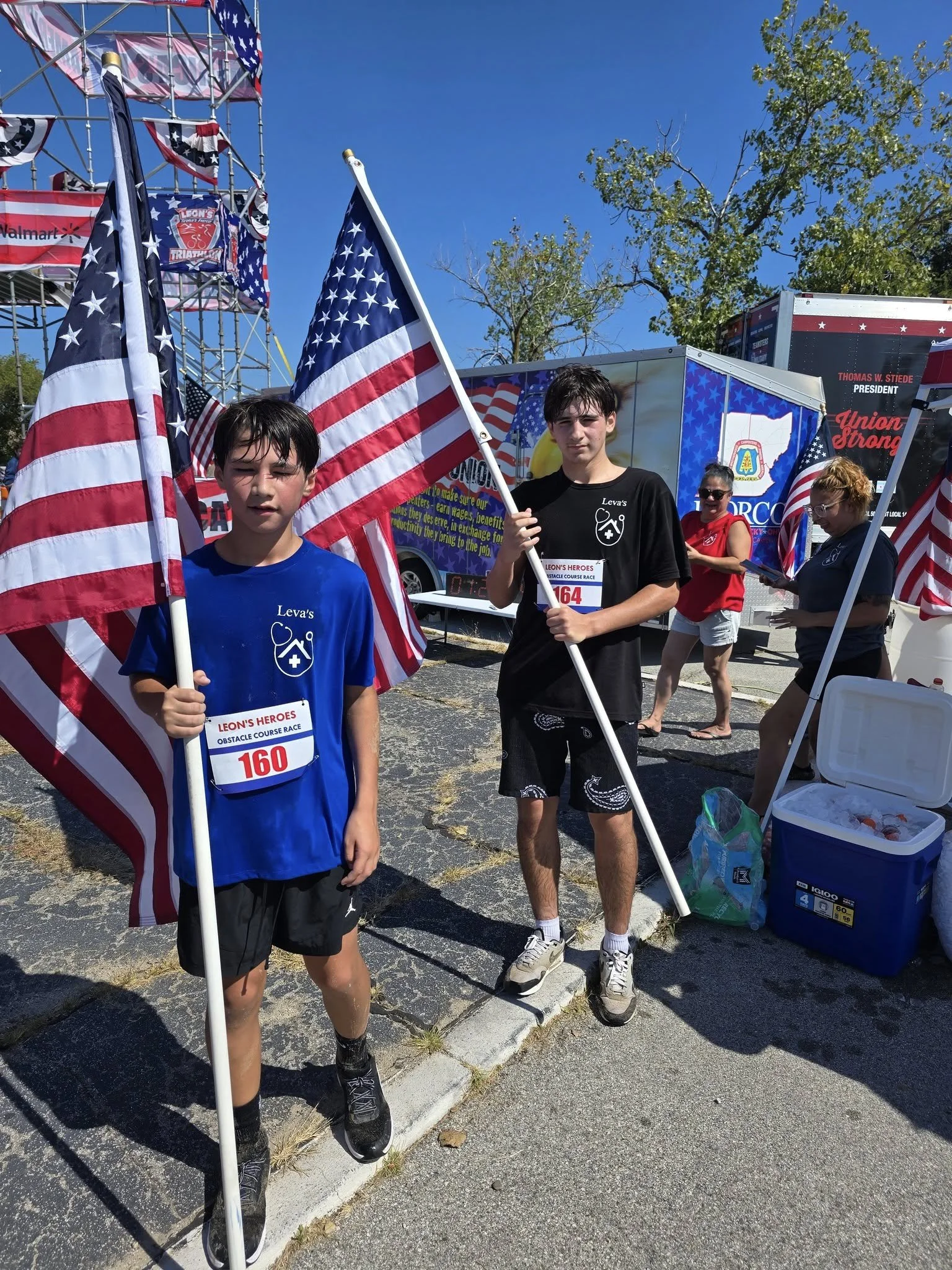 Two boys holding American flags at an outdoor event, with a crowd of people and event trucks in the background.