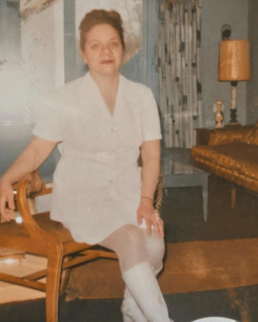 A woman dressed in a white outfit sitting on a wooden chair in a vintage living room.
