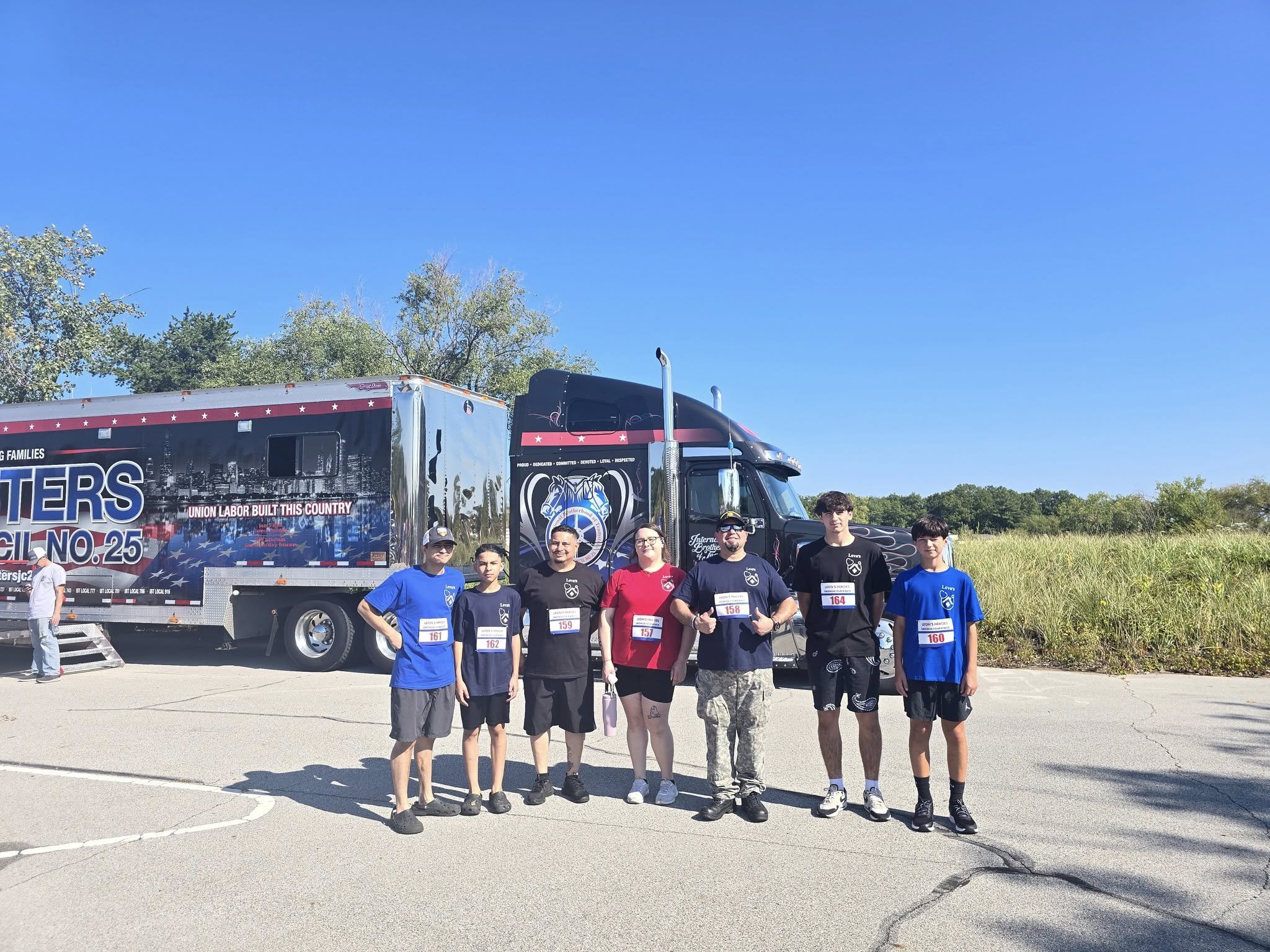 A group of seven people posing in front of a large black and red truck with a collage of images and text, including the words "Union Labor Built This Country" and "No. 25." They are standing on a paved area under a clear blue sky, with trees and grass in the background. Some are wearing sports jerseys with race bibs.