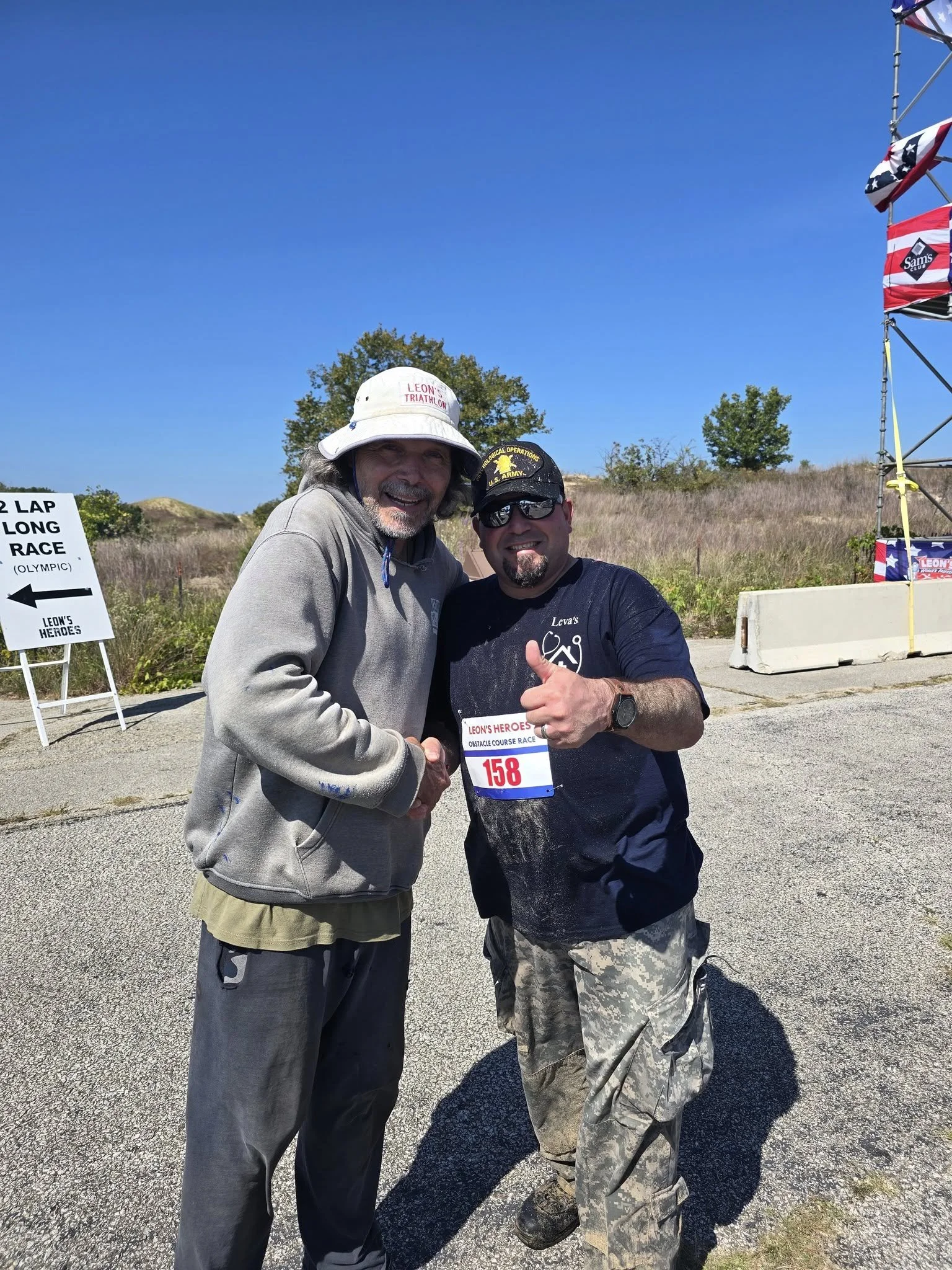 Two men smiling and shaking hands at an outdoor racing event on a bright, sunny day. One man is wearing a wide-brimmed hat and gray jacket, and the other is wearing sunglasses, a black cap, and camouflage pants. There is a sign in the background that says "2 Lap Long Race" and a race bib with the number 158.