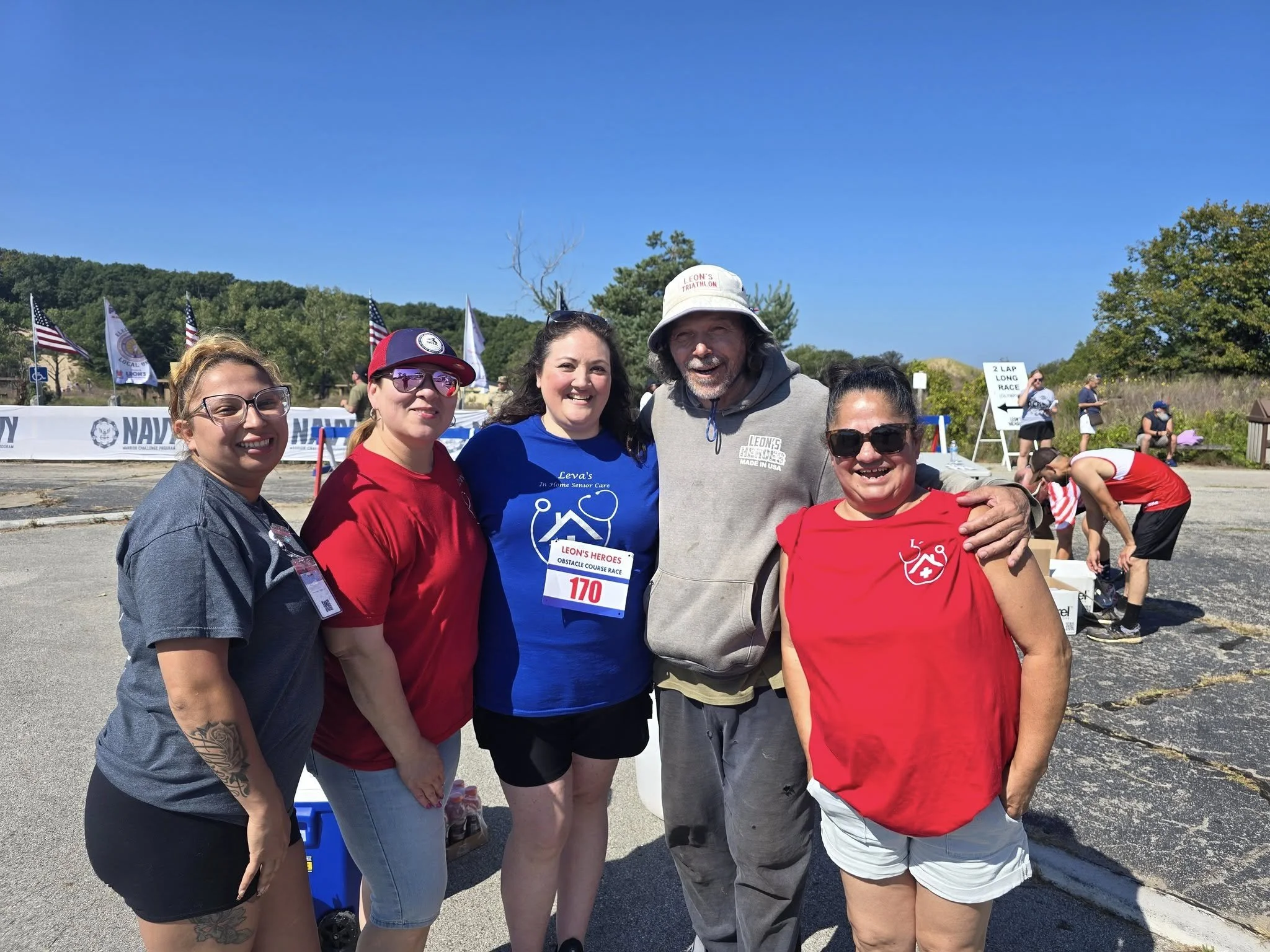 Group of five people smiling at an outdoor event, with flags and event signs in the background, during a sunny day.