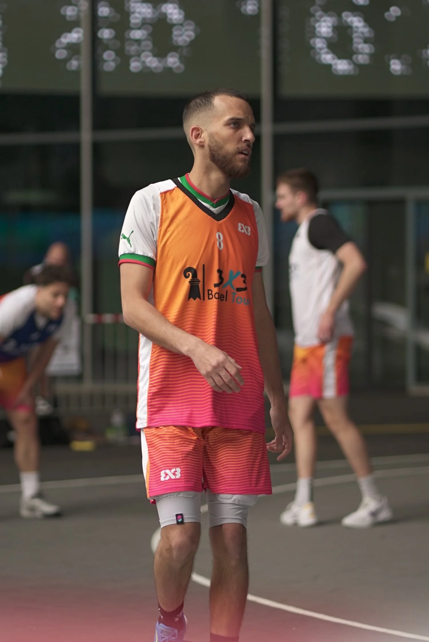 Basketballspieler in orange und pink Uniform auf einem Basketballfeld, im Hintergrund andere Spieler, Indoor-Sporthalle.