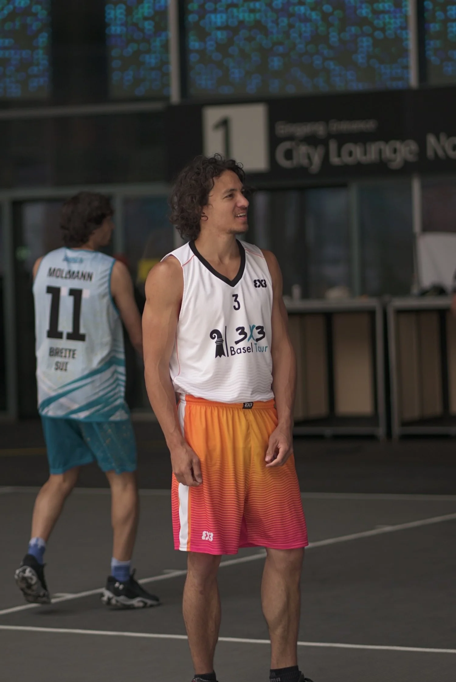 Zwei männliche Basketballspieler auf einem Indoor-Spielfeld, einer steht im Vordergrund und der andere im Hintergrund, beide tragen Turnschuhe und Sporthosen, einer trägt eine weiße Weste mit der Nummer 3 und der andere eine weiße Weste mit der Numme