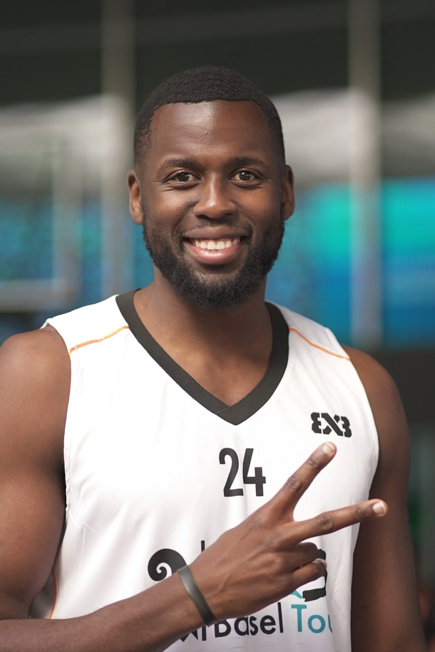 Ein lachender männlicher Basketballspieler in einem weißen Trikot, der die Nummer 24 trägt, zeigt mit zwei Fingern ein Peace-Zeichen.