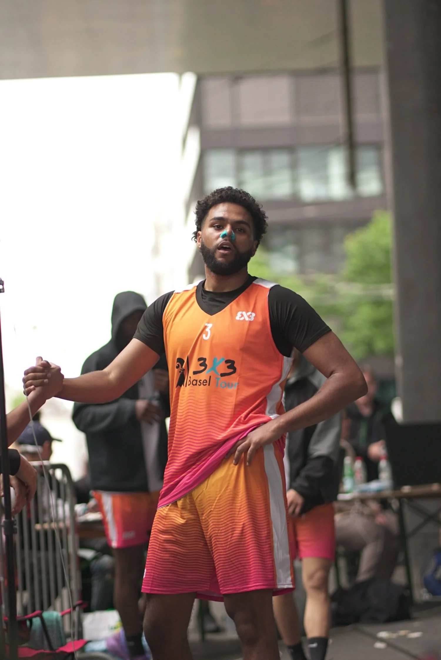Ein männlicher Basketballspieler trägt eine orange-rosa Weste mit der Nummer 3, während er nach einem Spiel in einer Spielhalle steht, umgeben von Menschen.