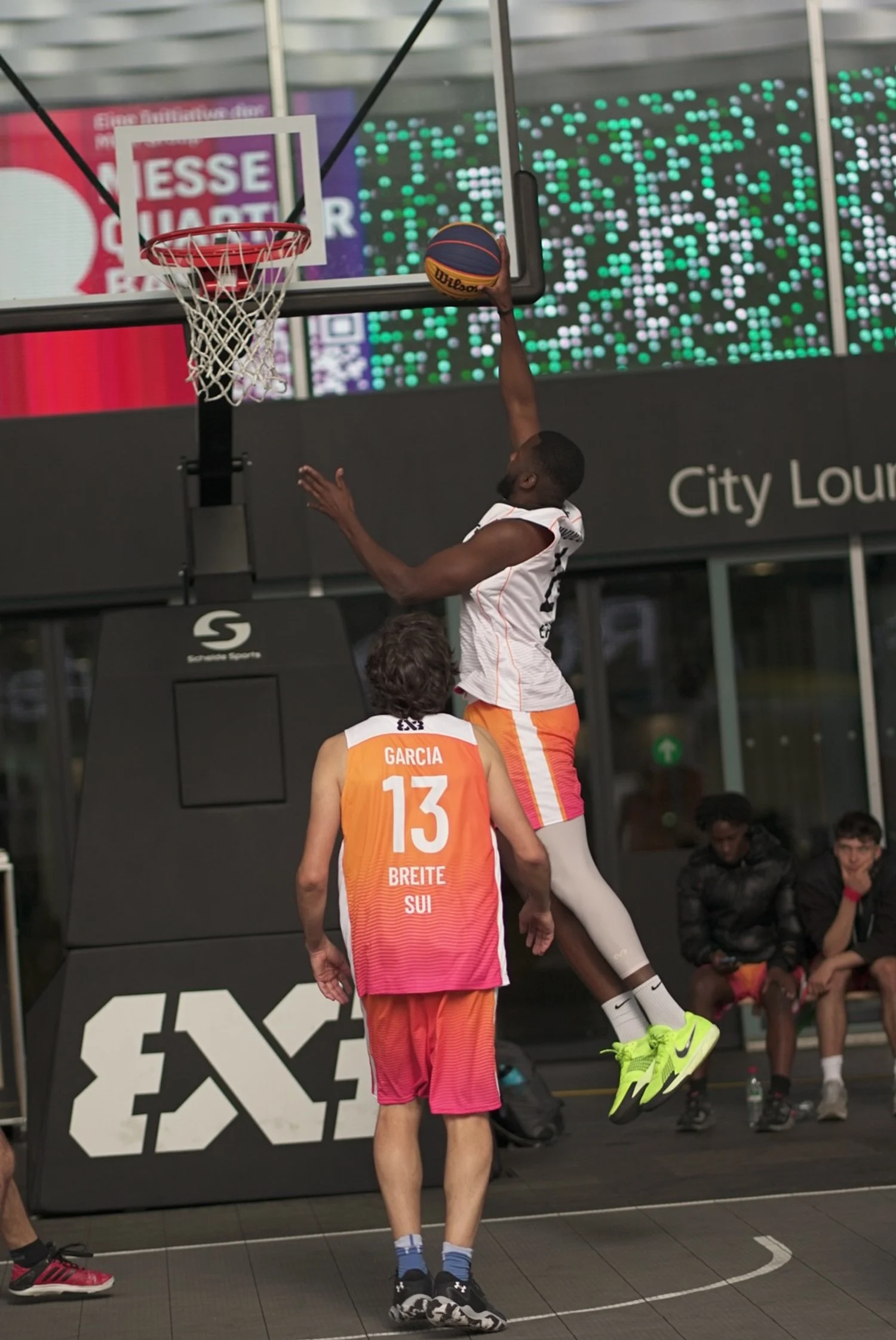 Ein Basketballspieler in den schwarzen Shorts springt zum Korb, während ein anderer in orangefarbenem Trikot mit der Nummer 13 unter ihm steht. Es ist eine Basketball-Action bei einem 3x3-Turnier, sichtbar durch das „3x3“-Logo auf der Basketball-Abde