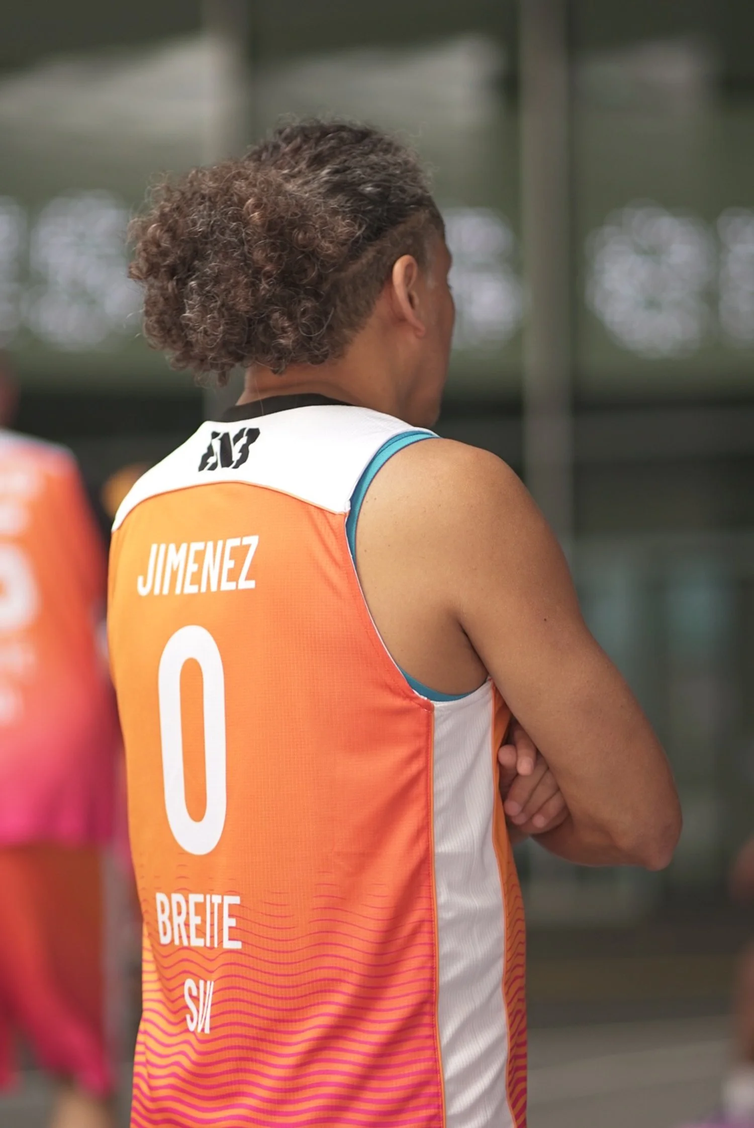 Basketballspielerin mit sichtbar aufgedrucktem Rückenpanorama, Name Jimenez und die Nummer 0, trägt ein orange-weißes Trikot.