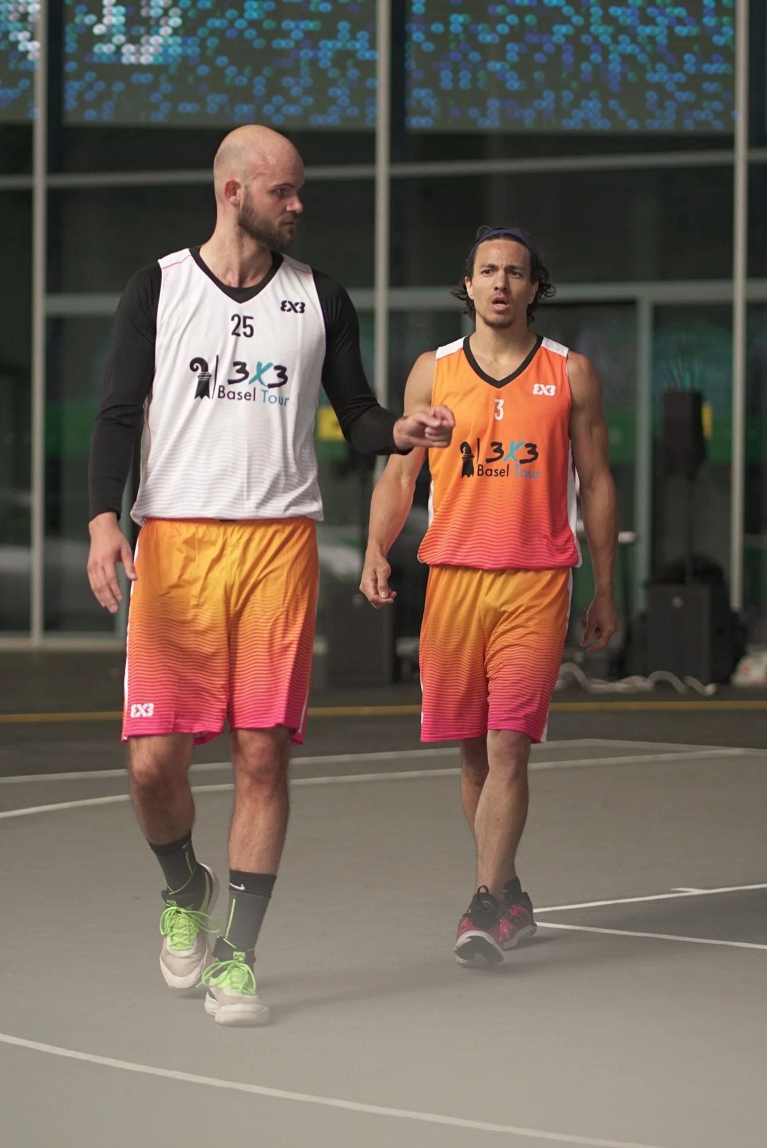 Zwei männliche Basketballspieler in bunten Trikots beim Gespräch auf einem Basketballfeld.