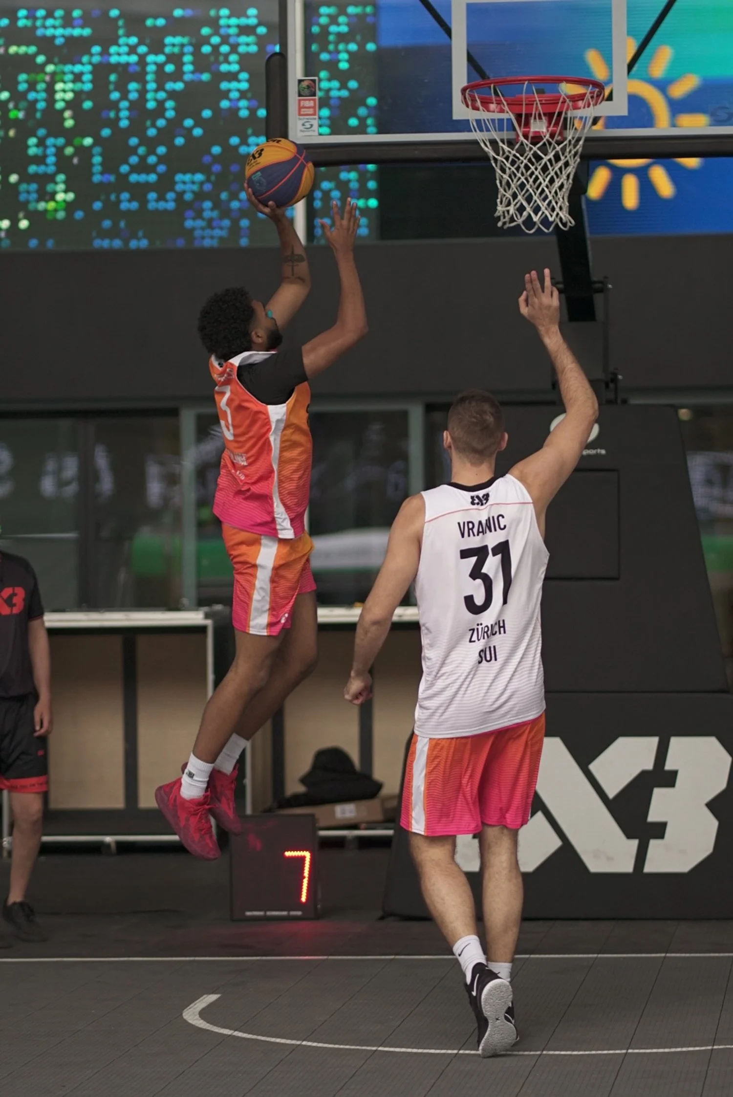 Zwei Basketballspieler im Spiel, einer springt zum Wurf. Der Spieler mit der Nummer 5 trägt ein rot-orangefarbenes Trikot, die Nummer 31 trägt ein weiß-rot-gestreiftes Trikot. Es ist ein Indoor-Basketballfeld, im Hintergrund stehen Anzeigen und eine