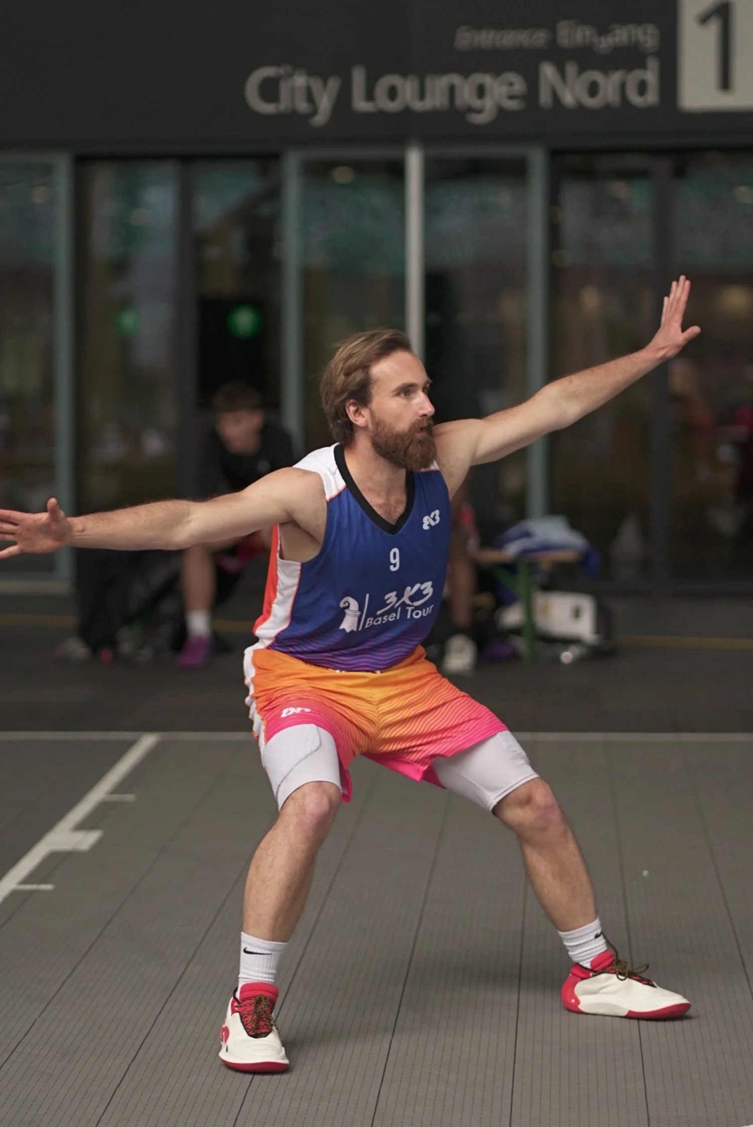 Ein Mann in bunten Basketball-Shorts und Sneakers macht eine Sportübung auf einem Parkettboden in einer Halle mit einem Schild im Hintergrund, das sagt 'City Lounge Nord'.