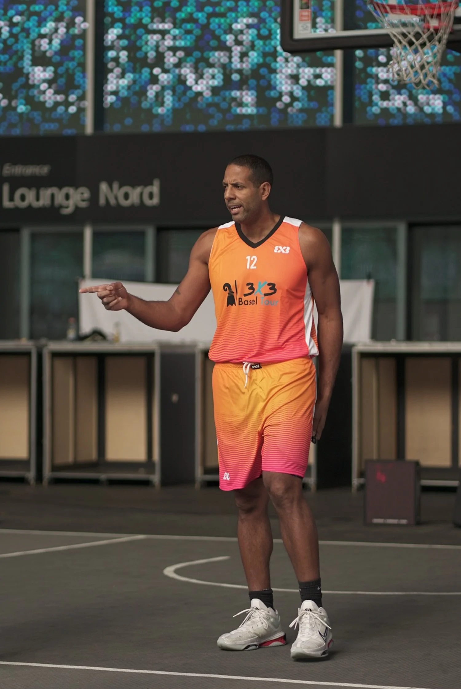 Ein männlicher Basketballspieler in orange-rosa Trikot und Shorts beim Training auf einem Basketballhof, im Hintergrund eine digitale Anzeige und das Schild "Lounge Nord".