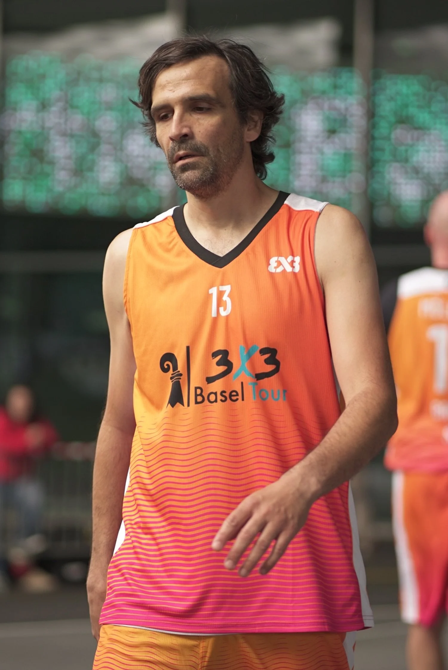 Ein männlicher Basketballspieler trägt ein orangefarbenes Trikot mit der Aufschrift '3x3 Basel Tour' und der Nummer 13, steht auf einem Basketballfeld.