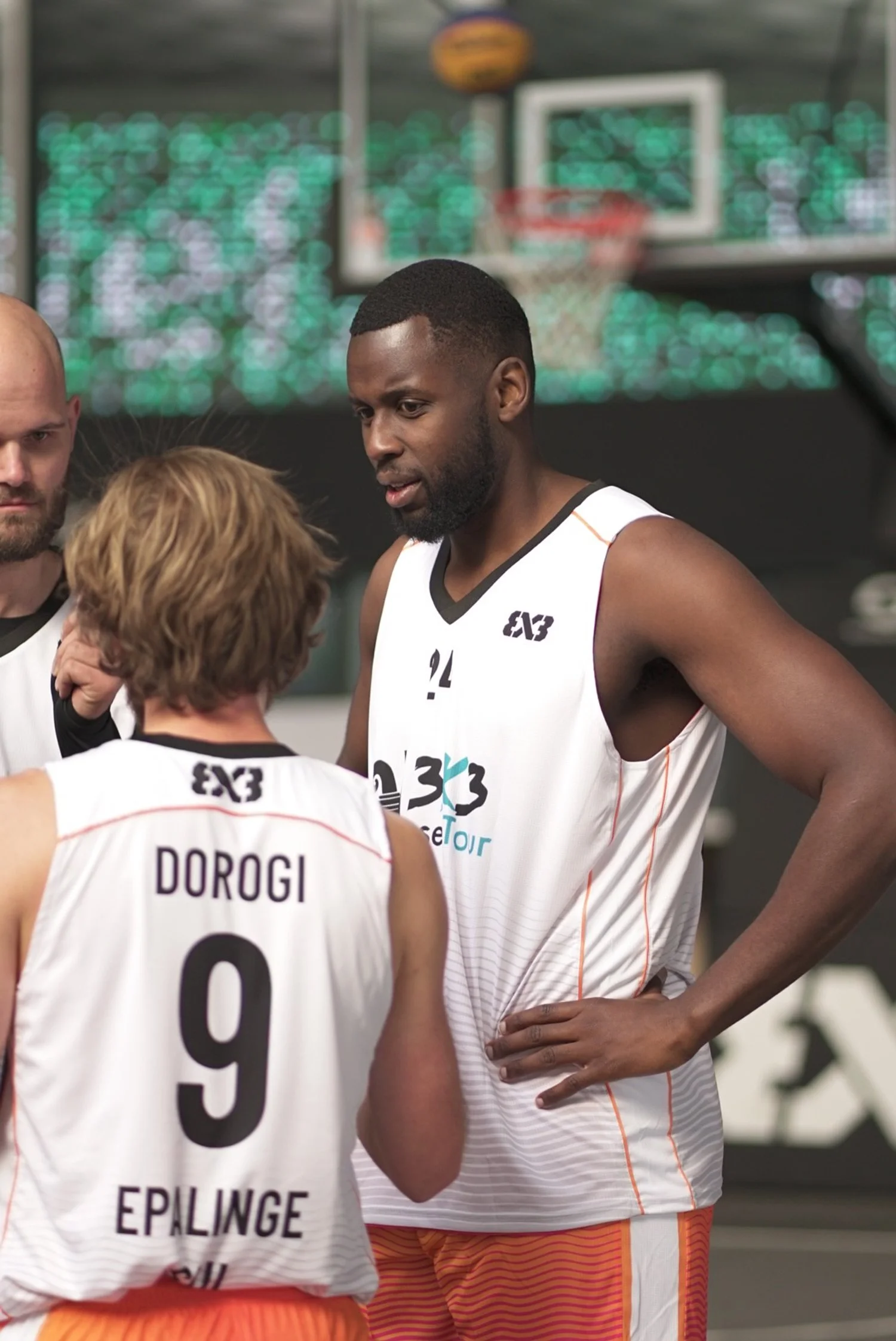 Zwei Basketbälle spielen, einer spricht mit einem Trainer, im Hintergrund Basketballkorb und bunte Lichter.