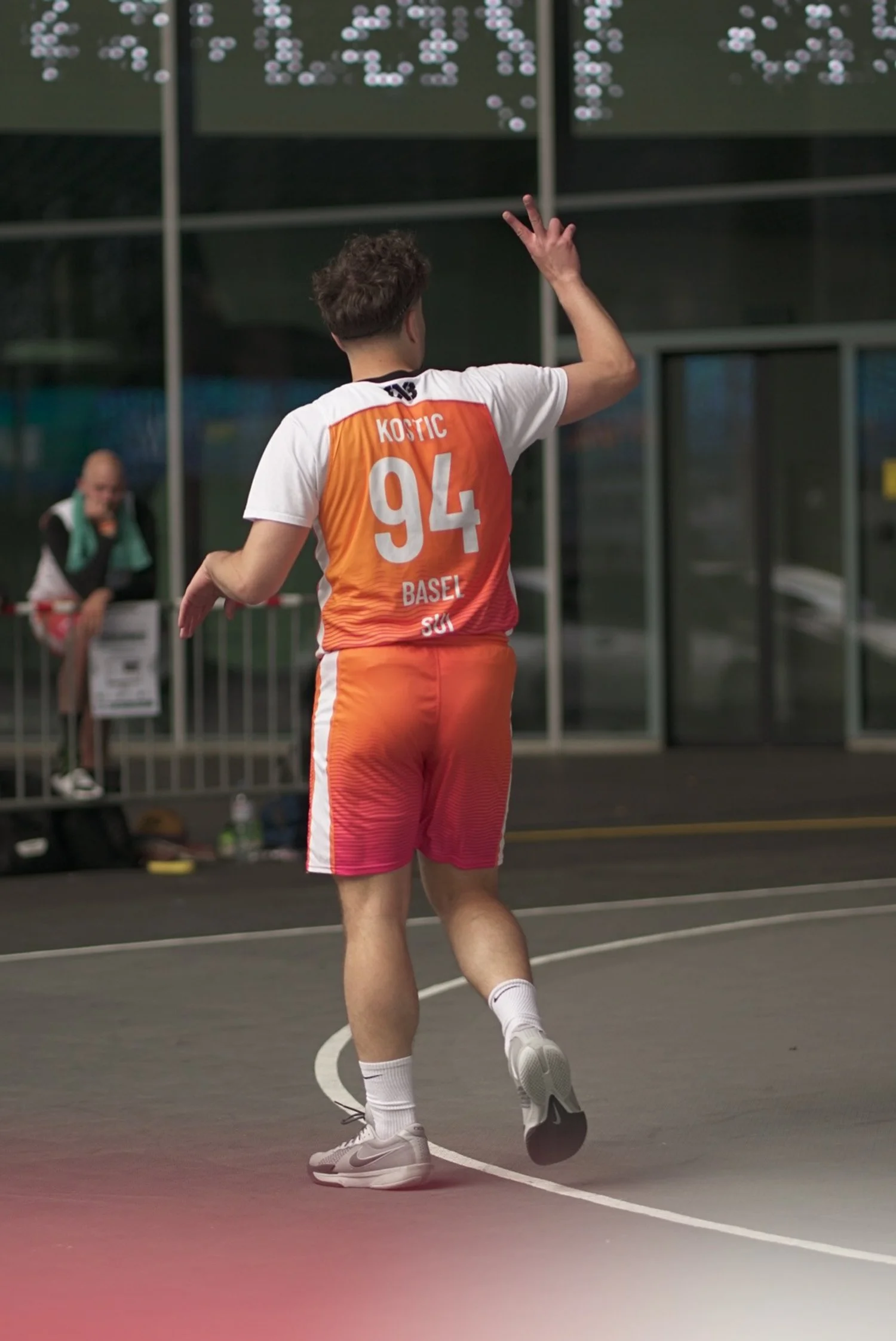 Ein Basketballspieler mit der Nummer 94 in einem orange-weißen Trikot auf einem Basketballfeld, der sich auf einen Freiwurf vorbereitet, während ein Assistent auf der Bank im Hintergrund sitzt.