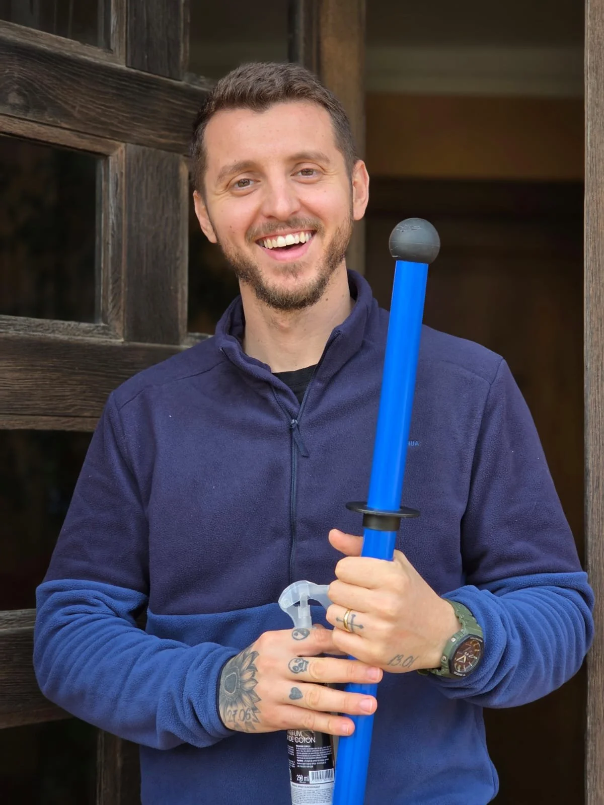 Un homme souriant tenant une lance à eau bleue, portant une montre et plusieurs tatouages, debout devant une structure en bois.