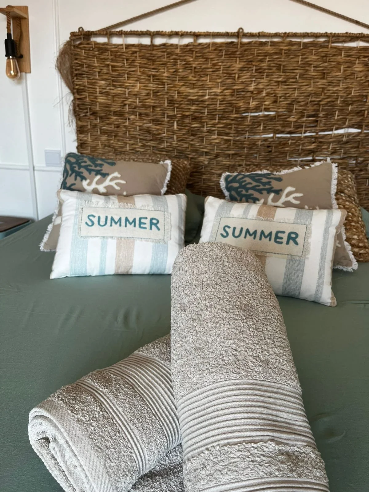Un lit avec un grand tête de lit en rotin, deux oreillers avec inscription "SUMMER" et deux autres avec un motif de corail, ainsi que deux serviettes enroulées de couleur beige claire.