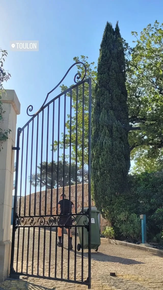 Une grille en fer forgé ouverte devant un chemin en pavés, avec un homme en vélo en arrière-plan et un grand arbre en forme de cône à côté, sous un ciel bleu.