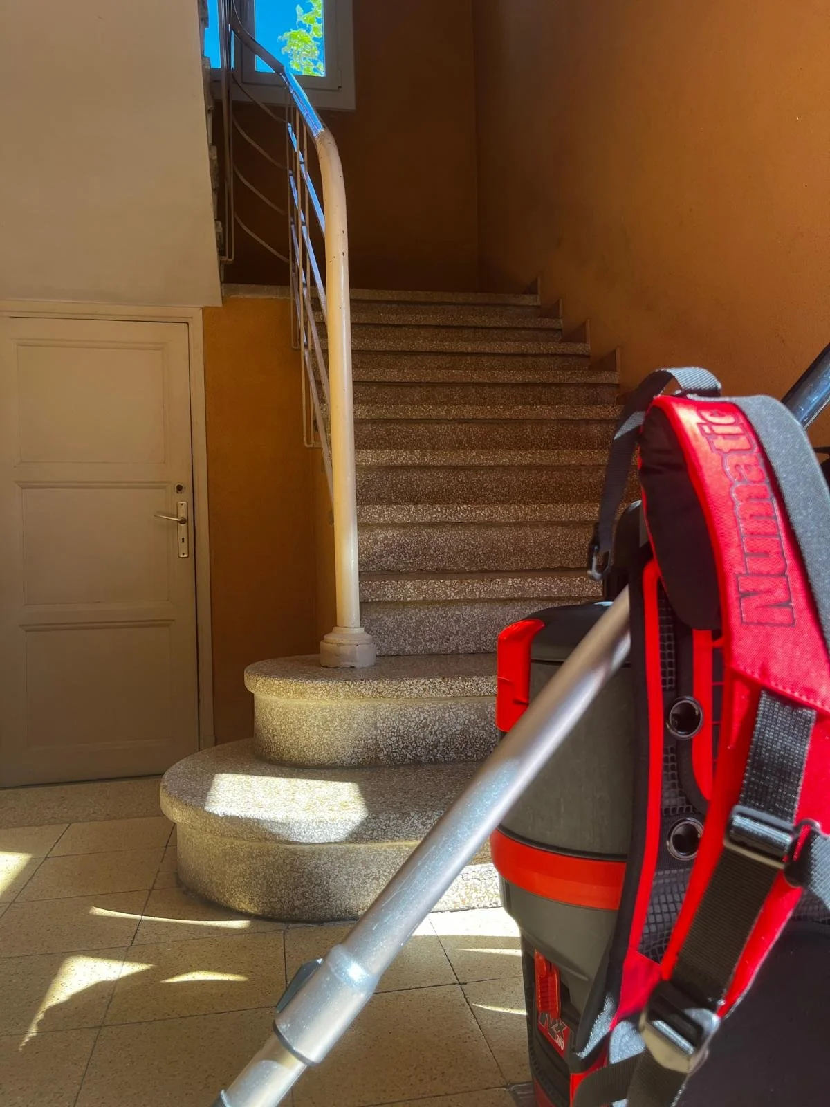 Escalier intérieur avec une rampe en métal, une porte et une fenêtre laissant entrer la lumière du soleil, avec un chariot de nettoyage à l'avant.