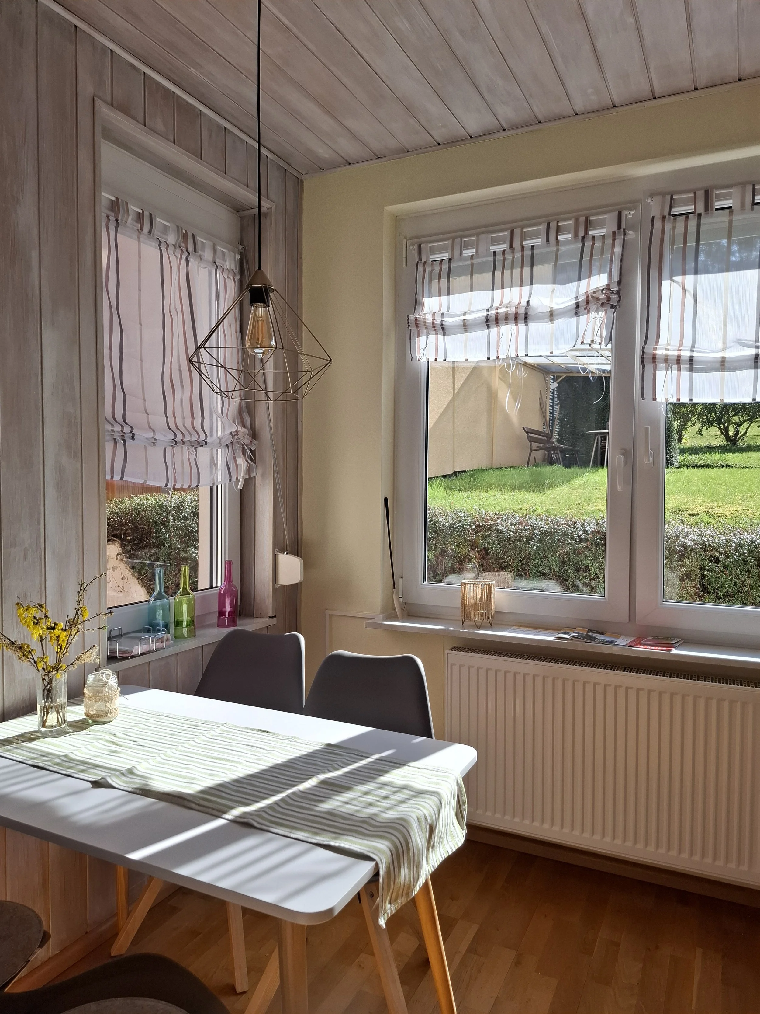 Helles Esszimmer mit Holzboden und weiß-grauem Tisch. Zwei moderne Stühle stehen am Tisch. Fenster mit gestreiften Rollos und Blick auf einen grünen Garten. Hängelampe mit Drahtgestell. Blumen und Deko auf dem Tisch.