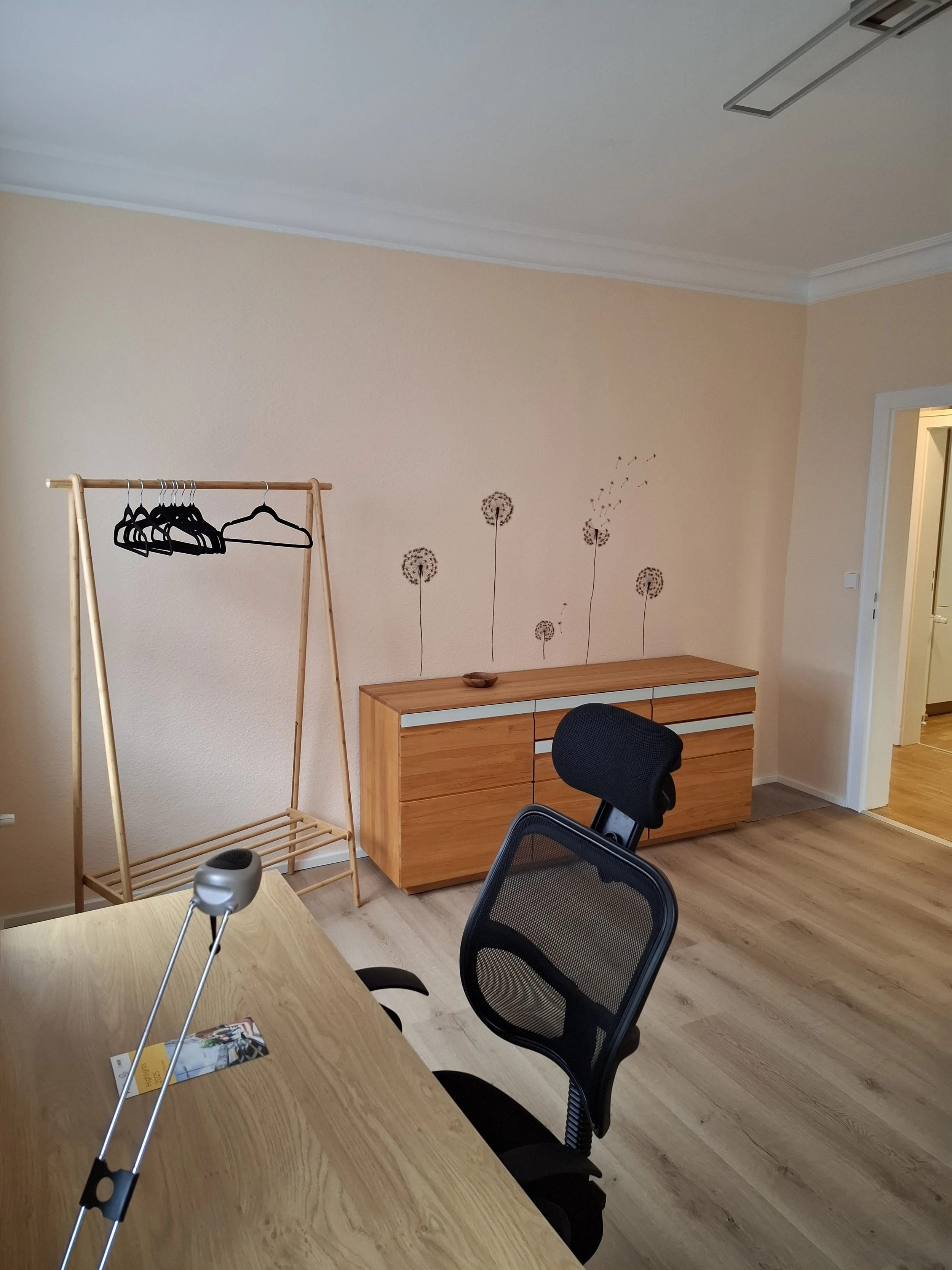 Innenansicht eines Zimmers mit einem Schreibtisch, einem schwarzen Bürostuhl, einem Holz-Kleiderständer mit mehreren Kleiderbügeln und einer Kommode im Hintergrund. An der Wand befinden sich dekorative Blumendekors.