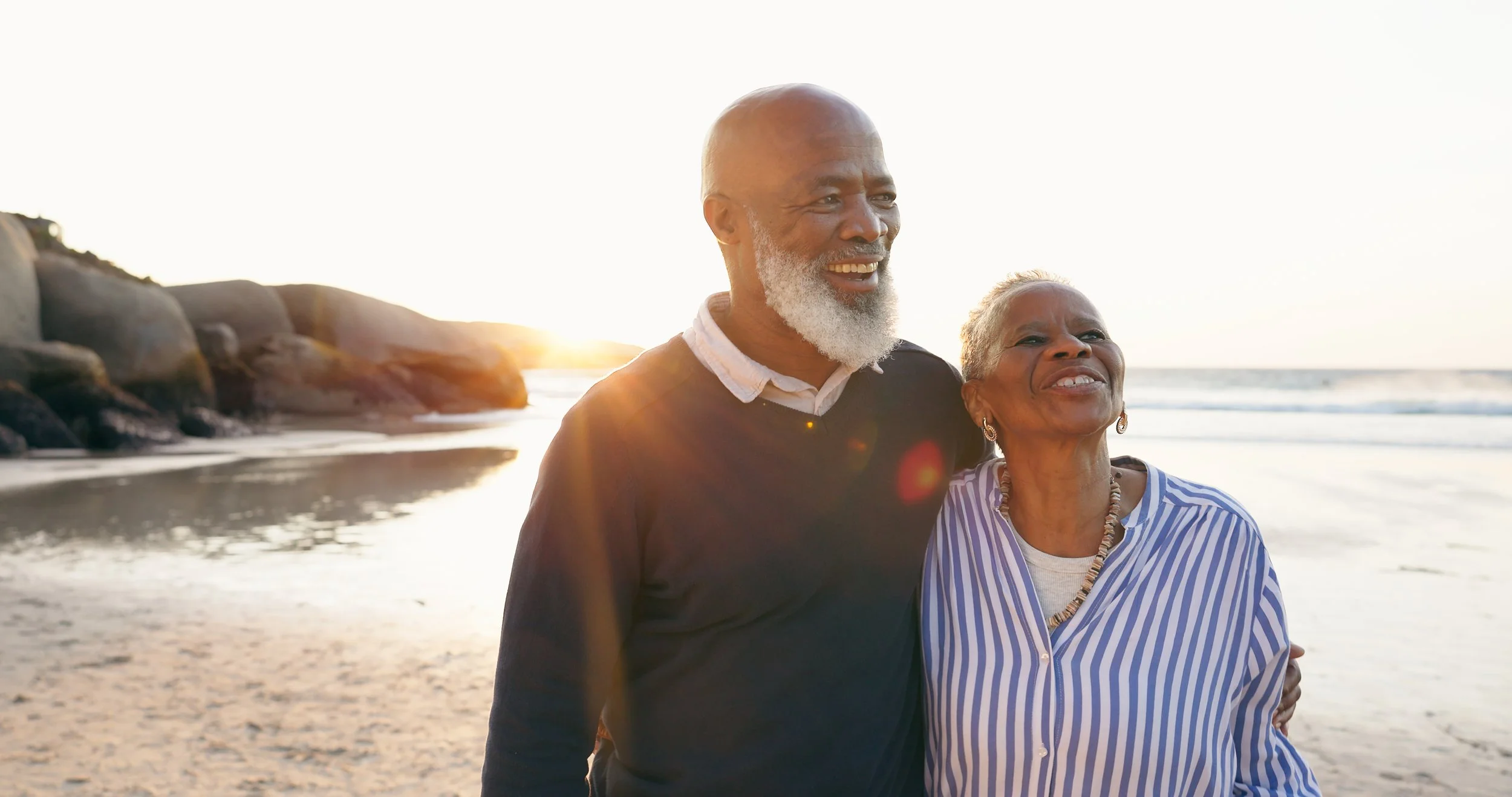 An elderly couple enjoying a walk on a beach at sunset, smiling and embracing each other.