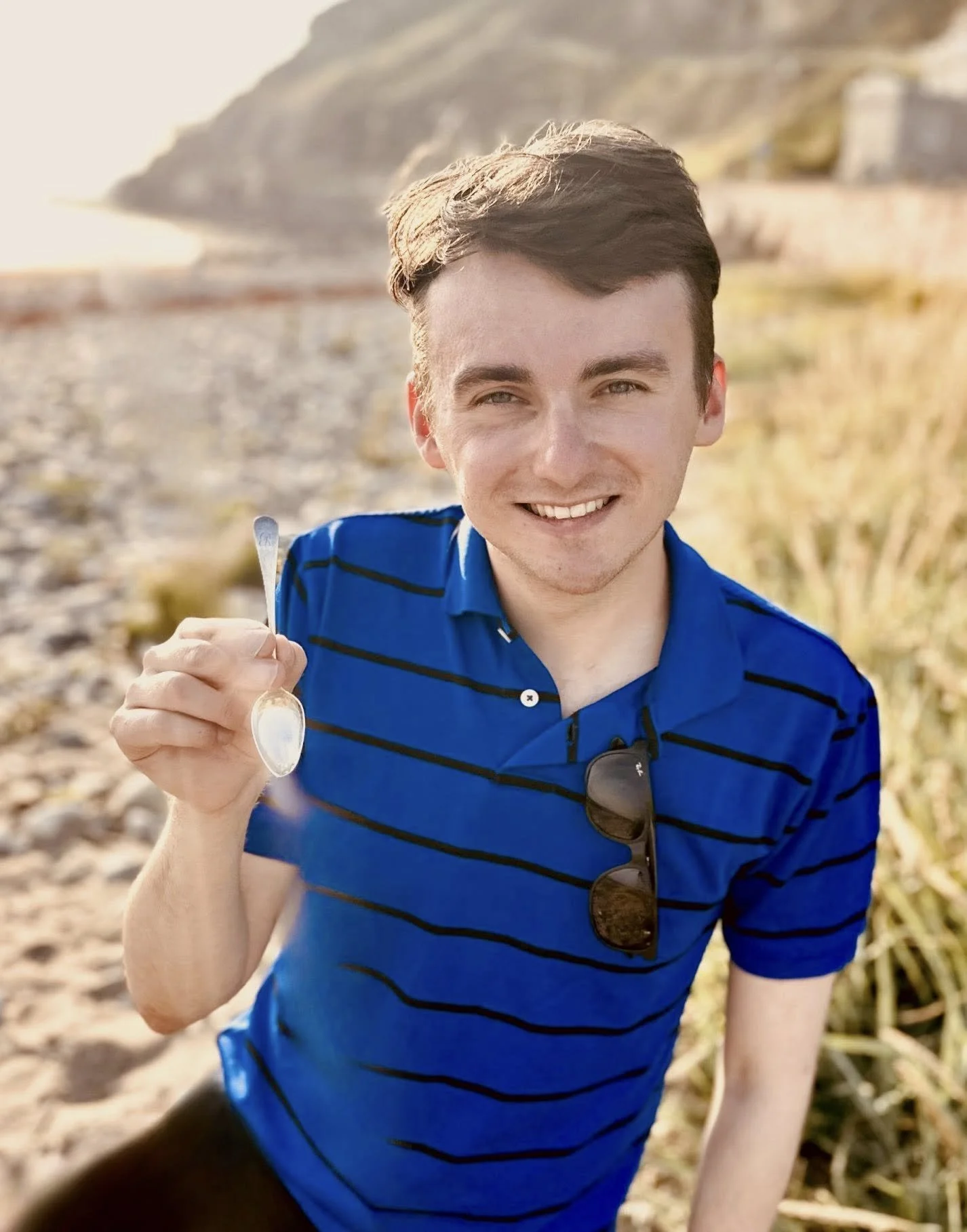 George wearing a blue striped polo shirt holds a silver spoon towards the camera outdoors at the beach with dunes and grass in the background.