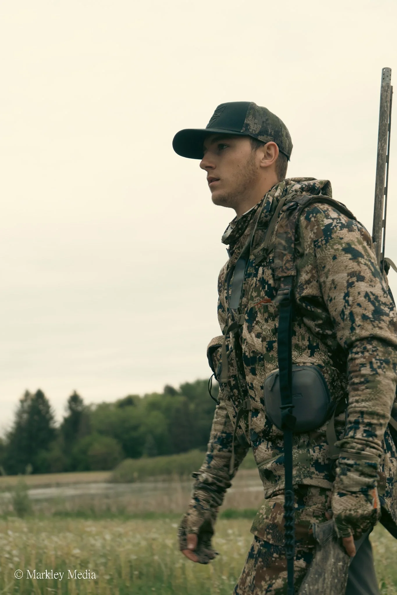Young man in camouflage clothing with a baseball cap, hiking outdoors near a river, carrying a backpack and gear.