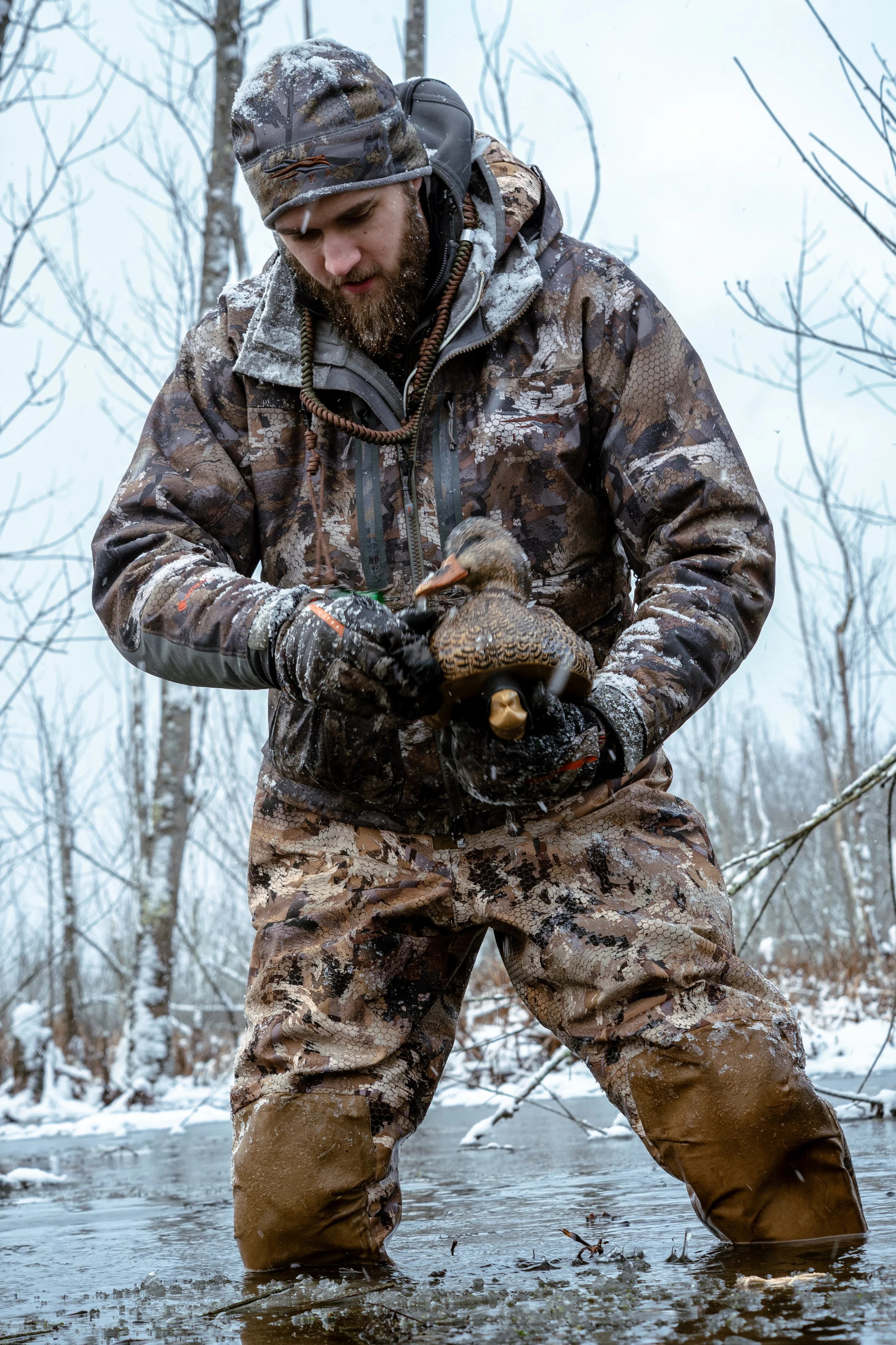 A man in camouflage winter clothing stands in a snowy stream, holding a duck he has just caught.