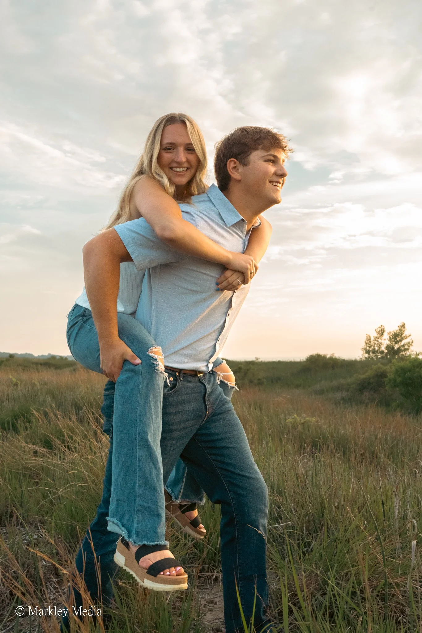 A young woman giving a piggyback ride to a young man in an open field during sunset, smiling and enjoying the moment.