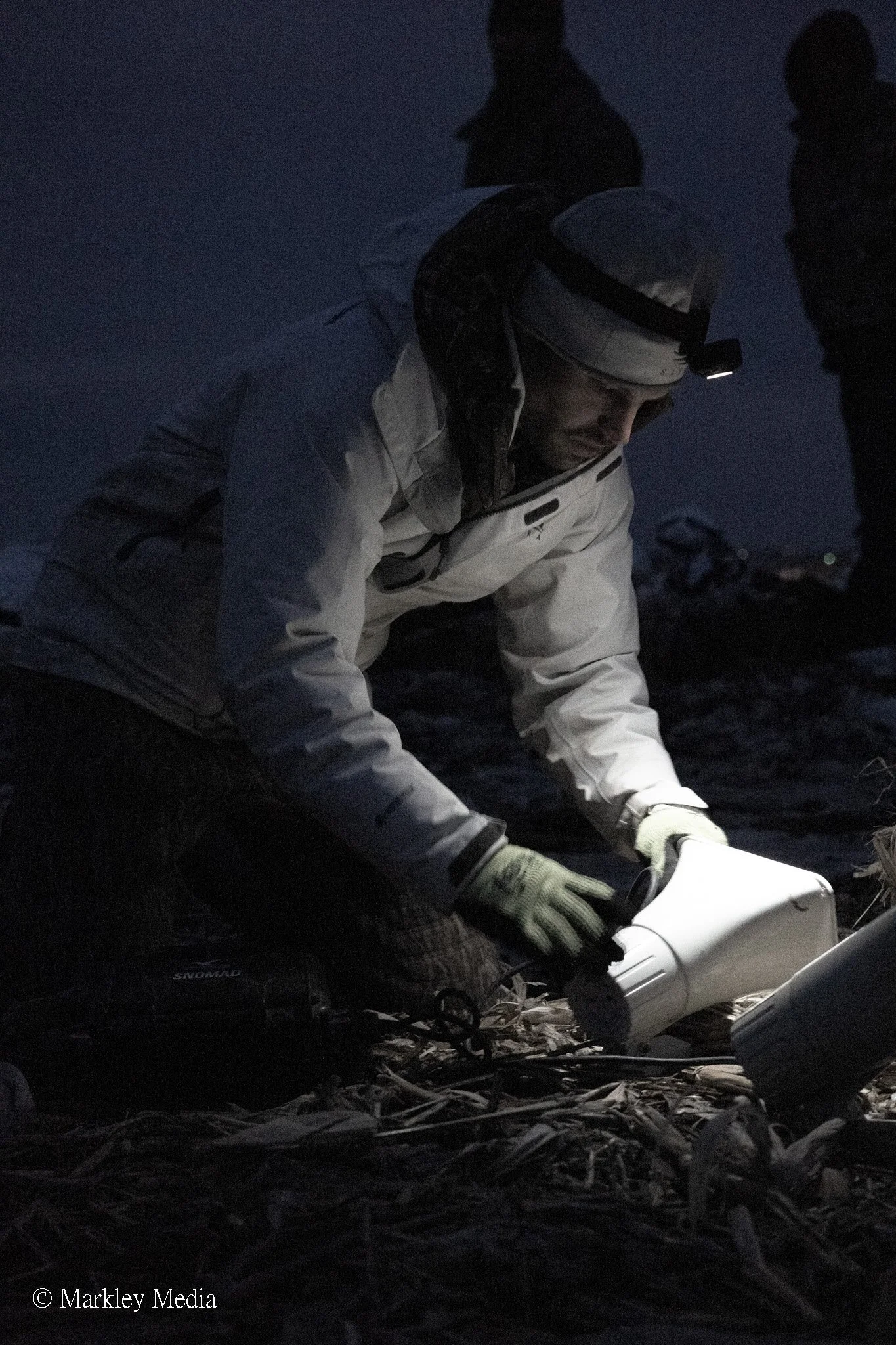 A person wearing a white jacket with a headlamp gathers samples in a dark outdoor setting, with other figures faintly visible in the background.