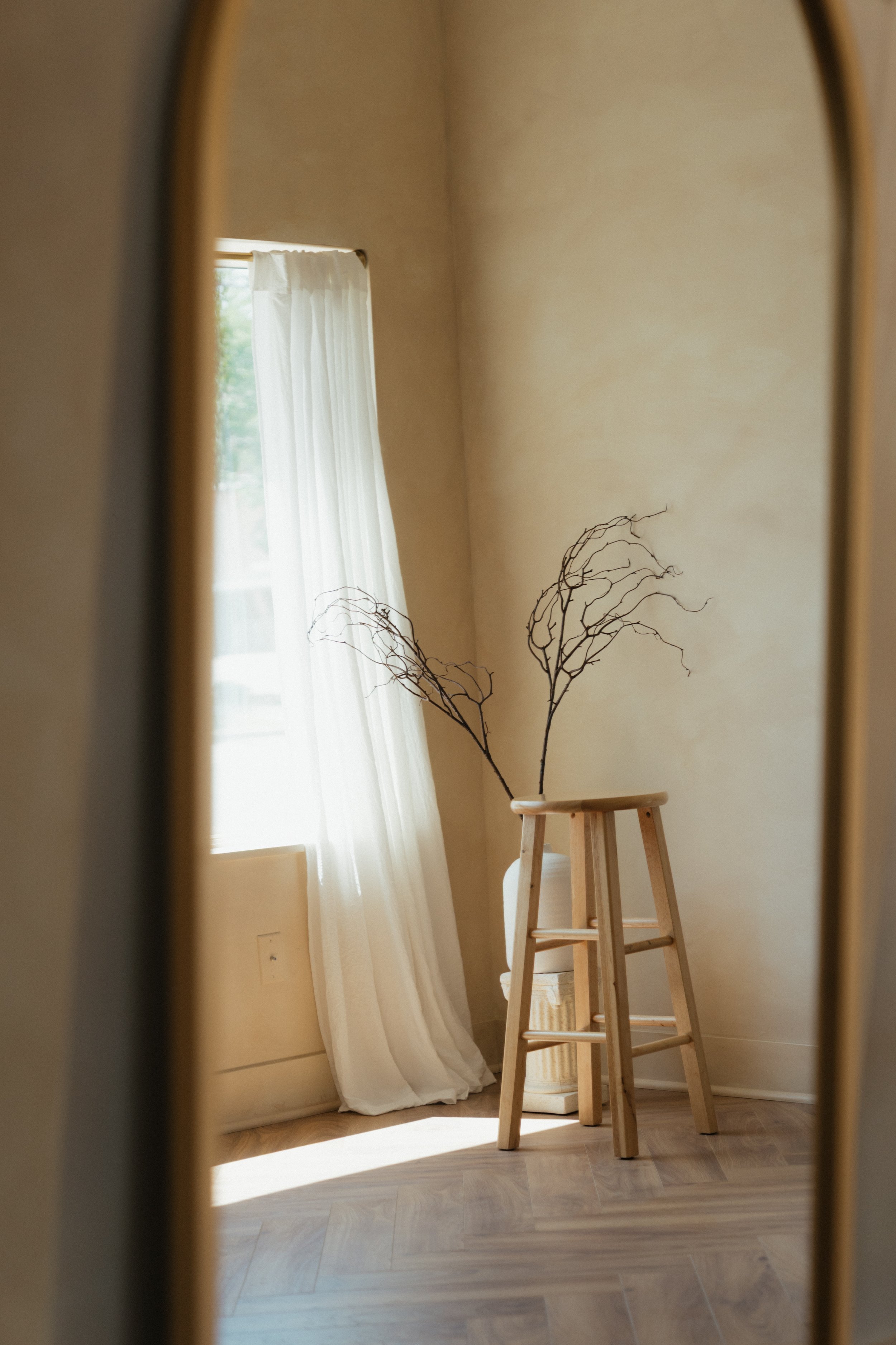 A mirror reflects a corner of a room with a wooden stool, dried branches in a vase, cream-colored walls, and a window with sheer curtains allowing sunlight in a rentable creative photo studio in East Grand Rapids Michigan.