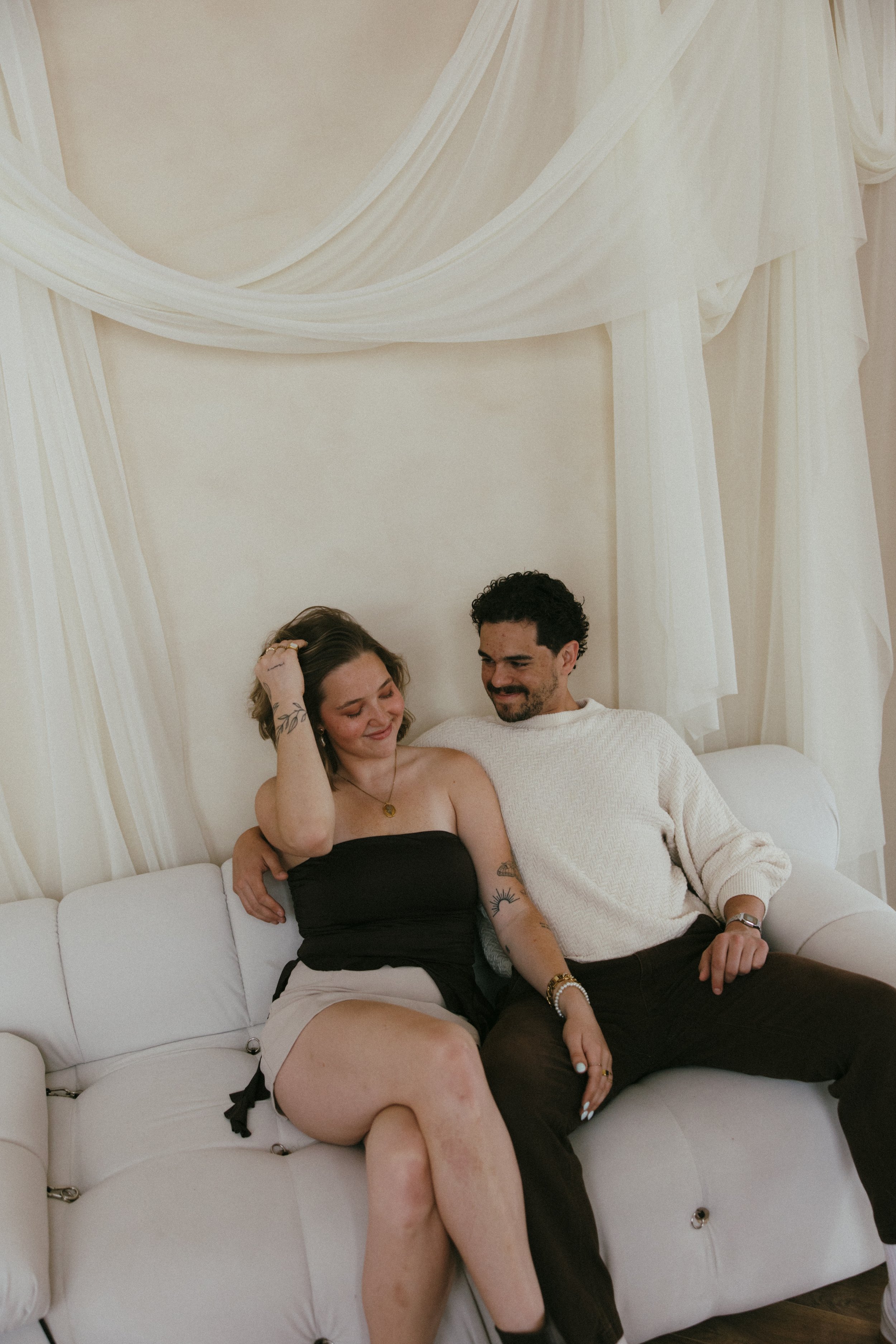 A man and woman sitting together on a white couch in a cozy room, smiling and looking at each other, with soft cream-colored drapes in the background in a rentable creative photo studio in East Grand Rapids Michigan.