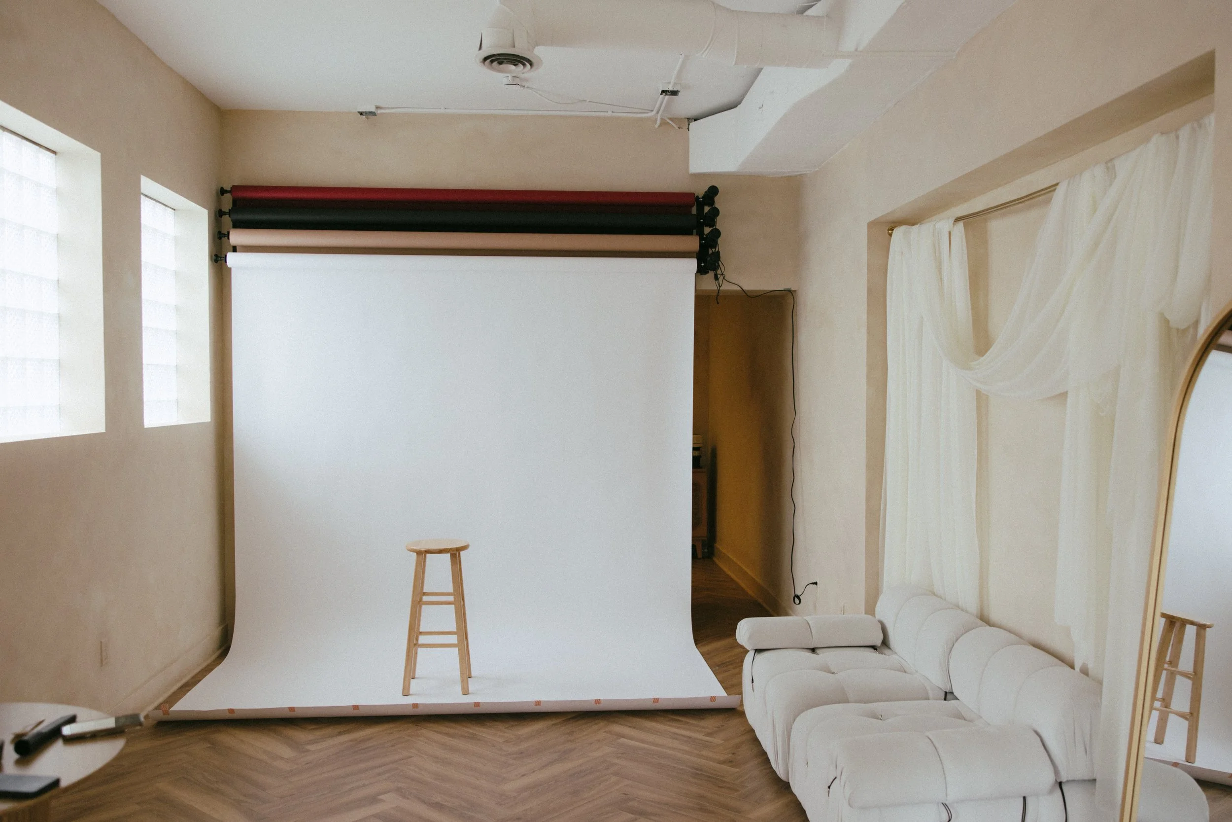 Indoor photo of a photography studio with a white backdrop, a wooden stool in front of it, and a white sofa to the right. There are windows with patterned glass and sheer curtains in a rentable creative photo studio in East Grand Rapids Michigan.