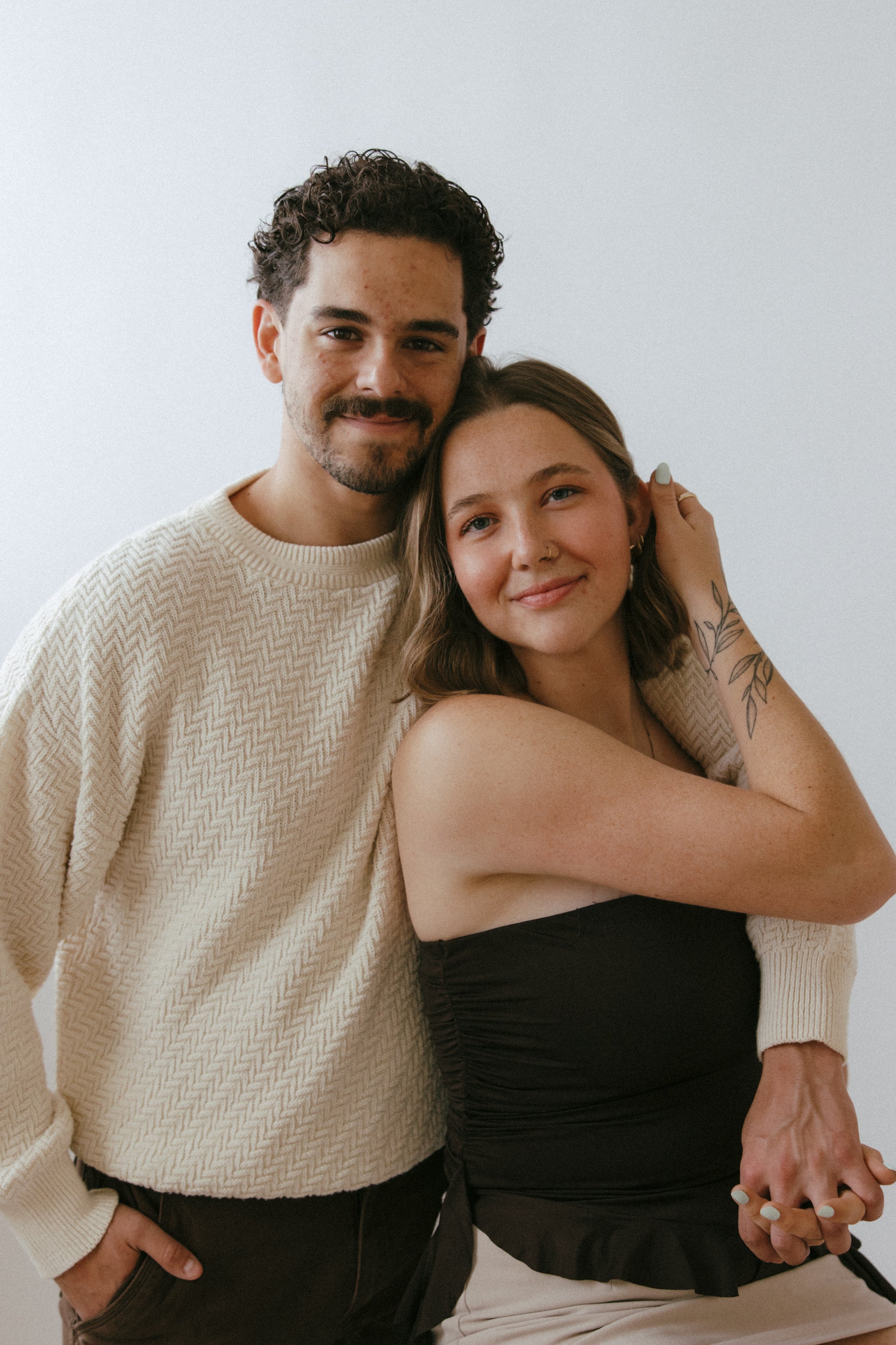A young man with curly dark hair and a beard, wearing a cream-colored sweater, standing next to a young woman with shoulder-length brown hair, a nose ring, and tattoos on her arm in a rentable creative photo studio in East Grand Rapids Michigan.