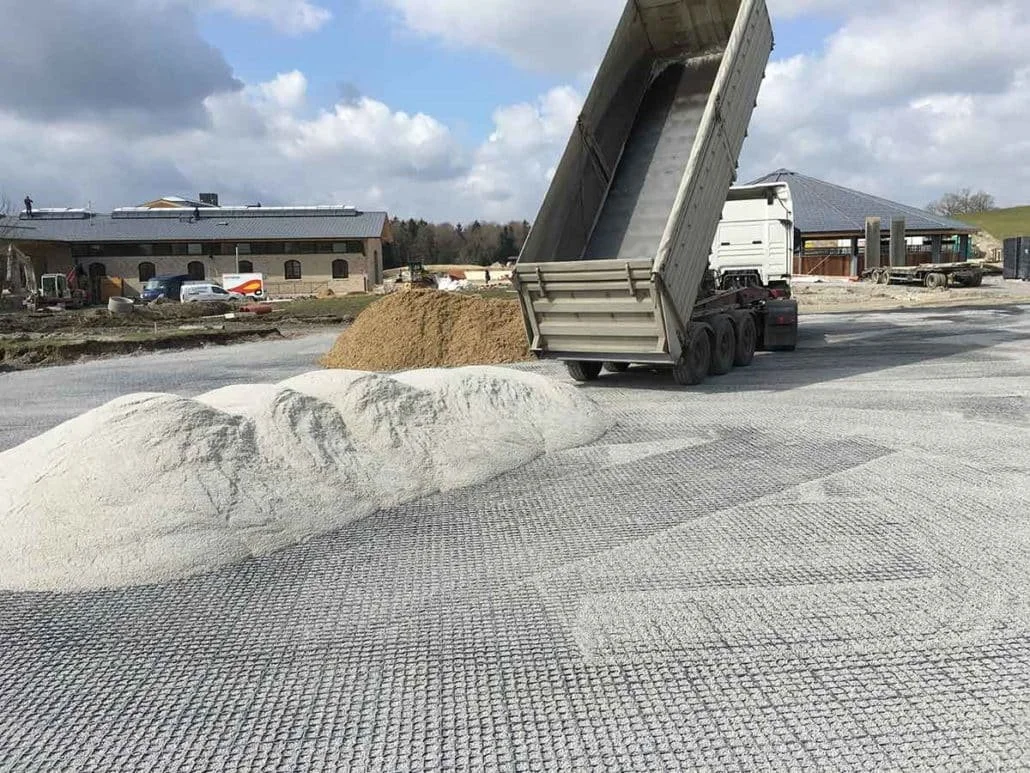 Dump truck unloading dirt and sand onto a construction site with a building in the background.