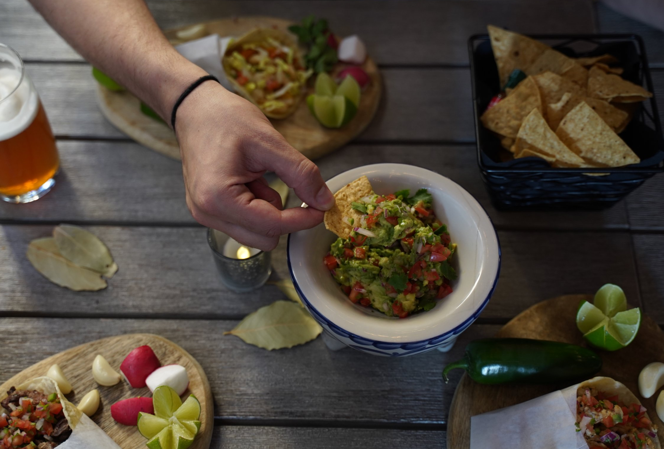 Person dipping a tortilla chip into a bowl of guacamole at a table with Mexican food items, including tacos, lime wedges, and a basket of chips, on a wooden surface.