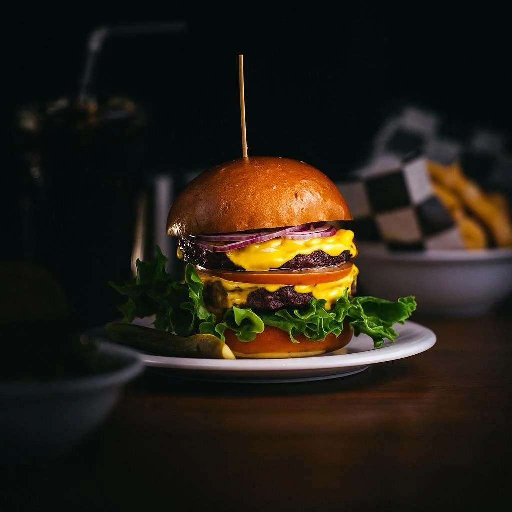 A gourmet cheeseburger with lettuce, tomato, cheese, onions, and beef patties, served on a plate with a pickle on the side.
