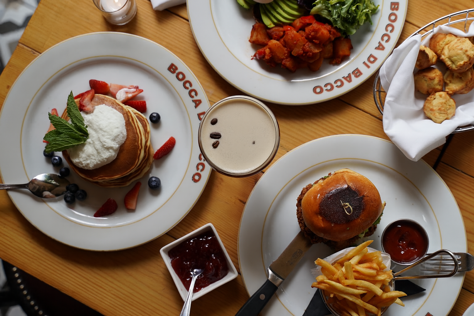 A table with various food dishes, including a stack of pancakes with strawberries, blueberries, whipped cream, and mint leaves, a sandwich with a bun, fries with ketchup, a salad with cucumbers and tomatoes, and fried snack items, along with a glass