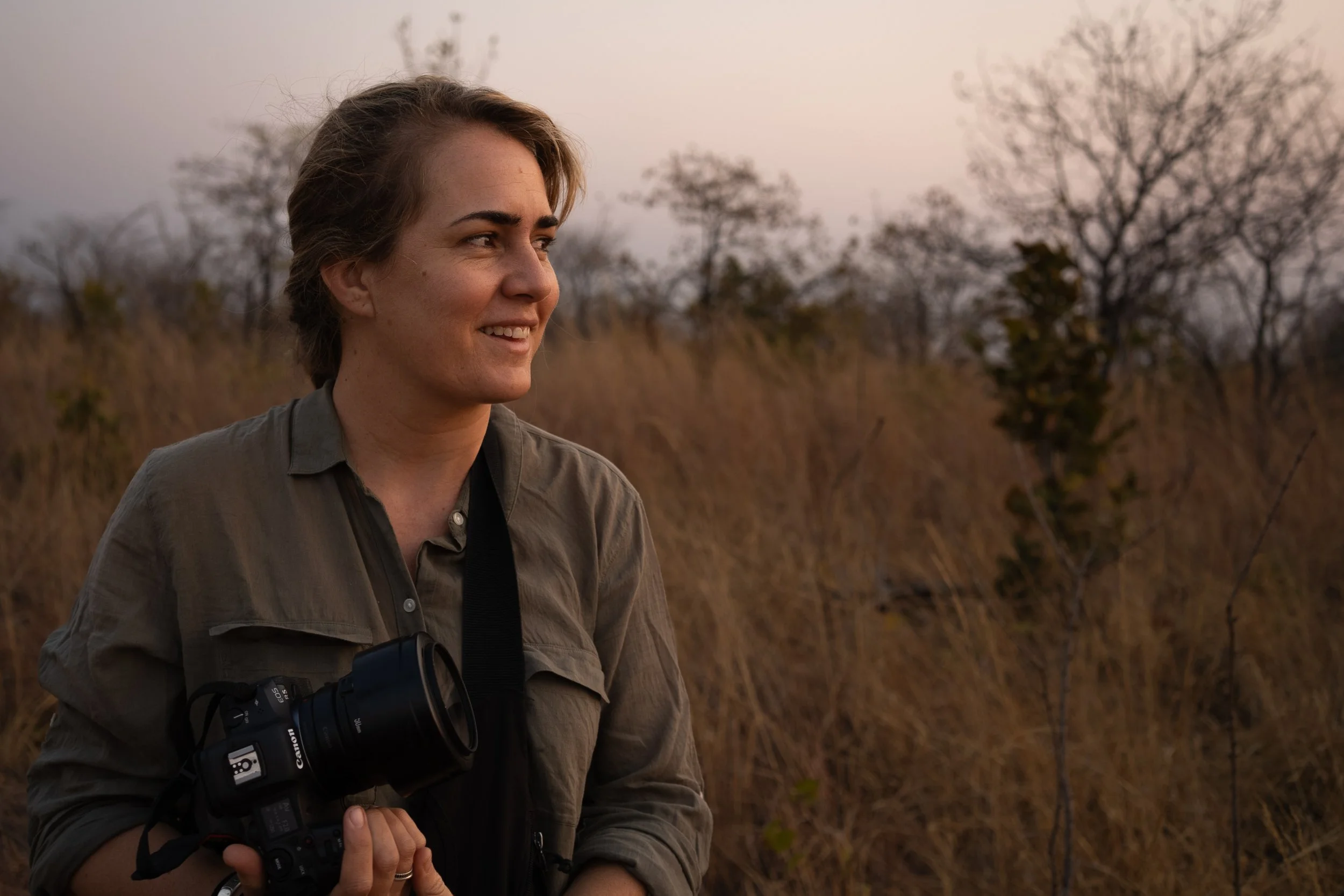 Mana Meadows holding a camera around her neck, standing outdoors in a natural setting with dry grass and leafless trees in the background at sunset.