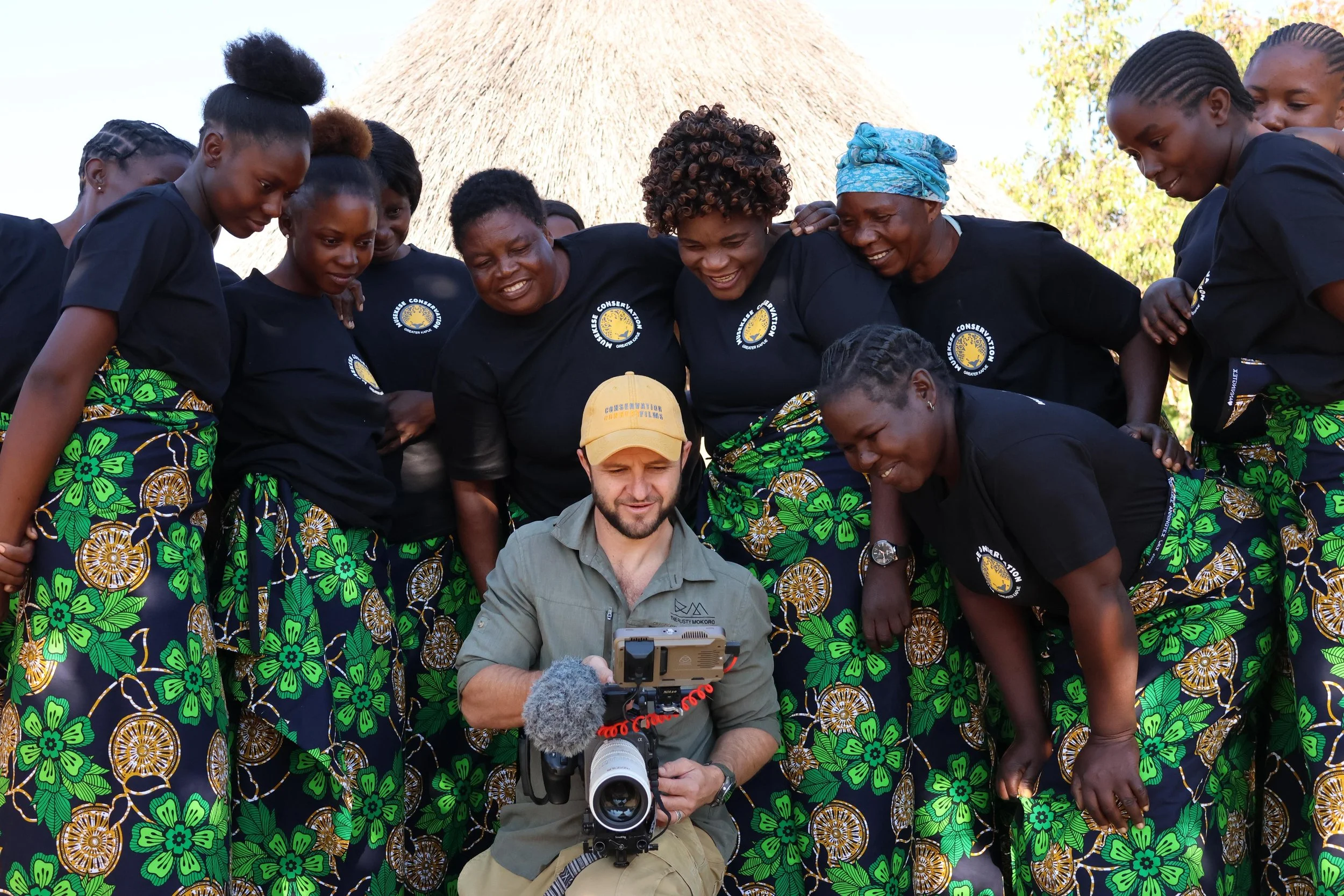 A group of women and a man gathered around, looking at a camera, outdoors in front of a thatched-roof hut. The women are dressed in matching black shirts and colorful skirts with green floral patterns, smiling and engaged. Matt Blair, conservation.