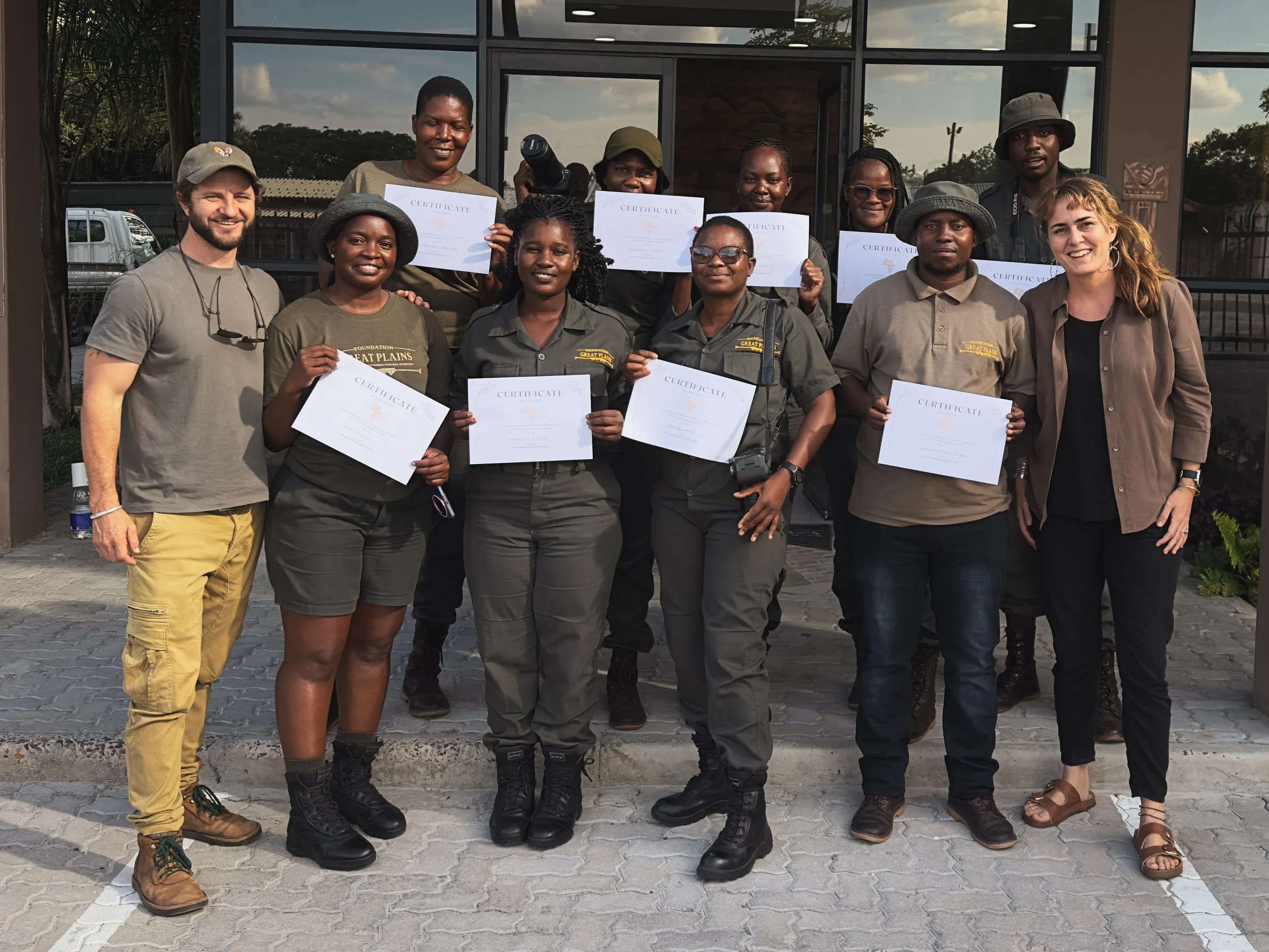 A group of ten people, including eight holding certificates, pose outside in front of a building. They appear to have completed a training or workshop, with some wearing outdoor or safari-style clothing and boots. The group looks happy and proud.