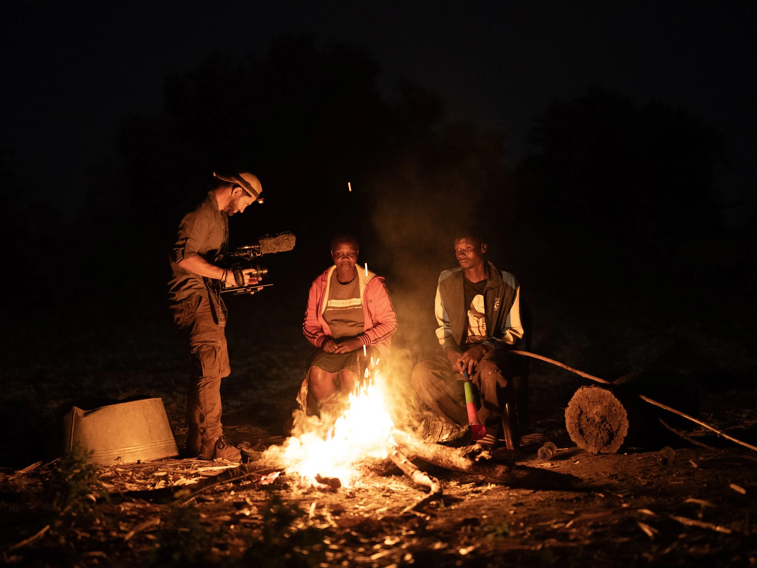 Two people sitting by a campfire at night, with a person filming them using a camera, and trees in the background.