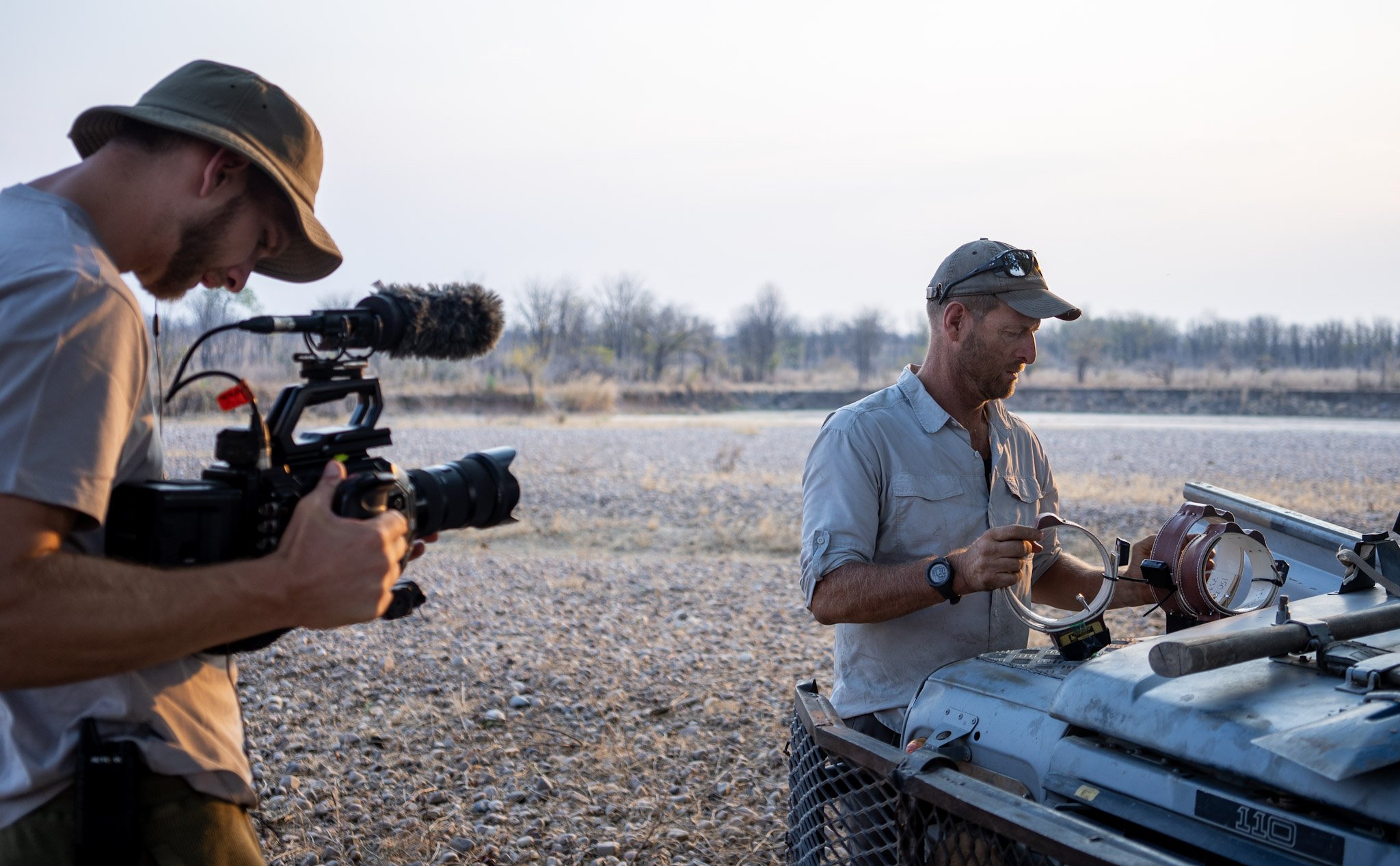 Rich Watson and Matt Becker filming for the conservation documentary, Abashimba. Two men outdoors near a body of water, one holding a camera and the other adjusting equipment on a vehicle, during daylight