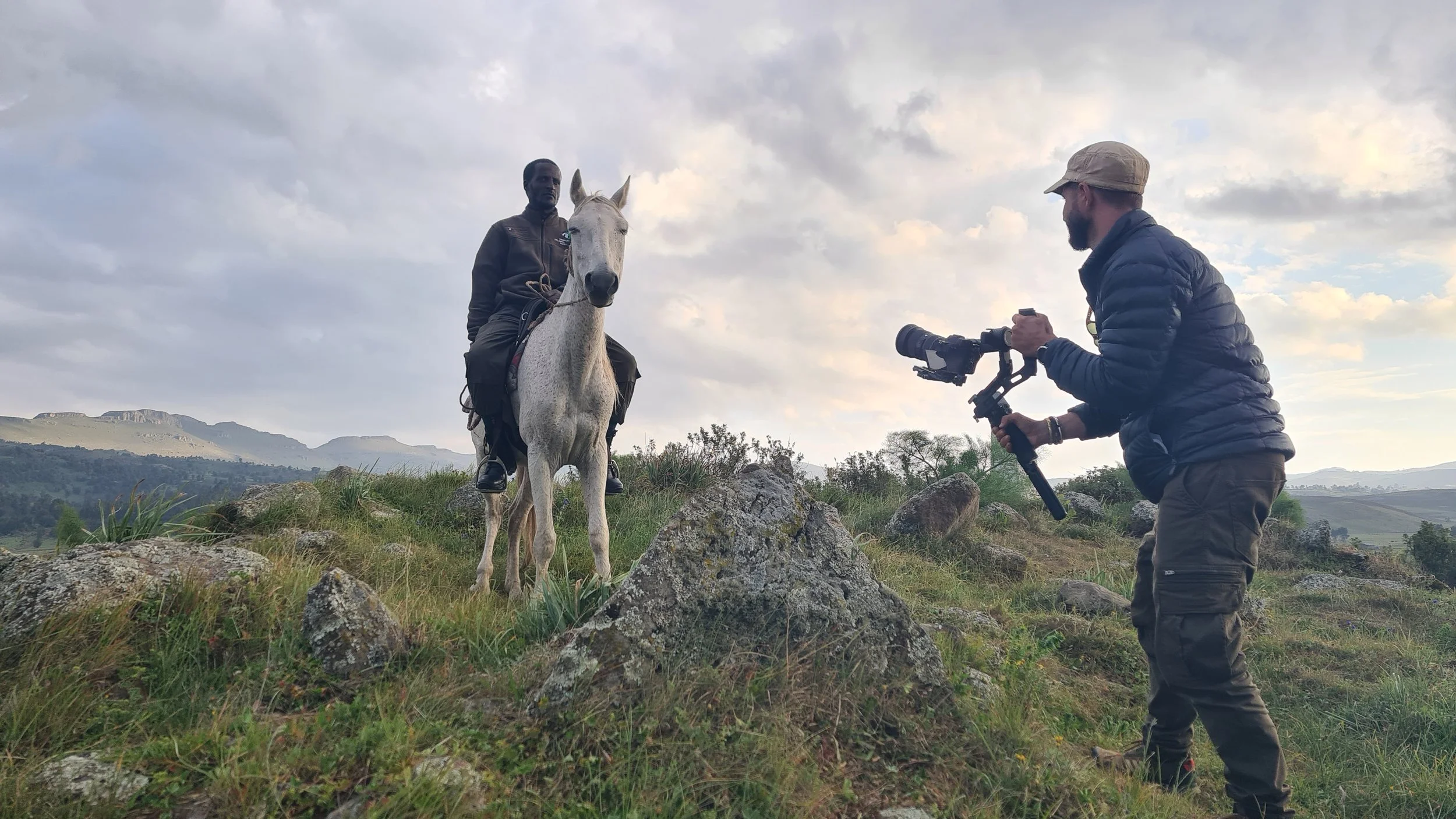 A man riding a white horse on a grassy hill with a scenic landscape in the background, while another man films him using a camera mounted on a stabilizer.