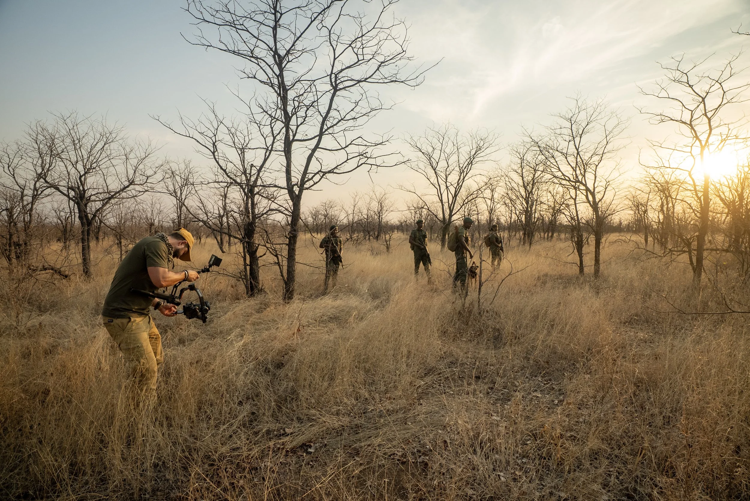 Five individuals, including Matt Blair conservation filmmaker with a camera, walking in a dry, leafless forest during sunset.