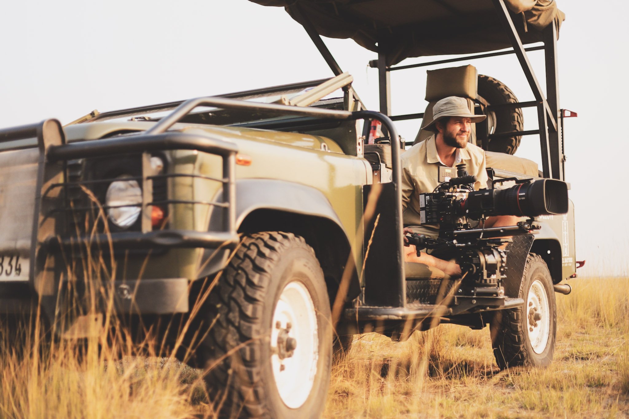 A man wearing a hat and outdoor clothing operating a professional camera mounted on a tripod, sitting in the open back of a safari-style vehicle in a grassy plain during sunset.